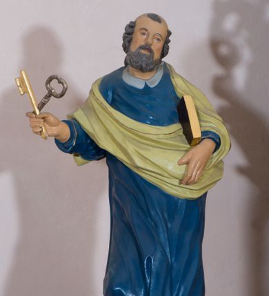 Zdjęcie nr 1: Rzeźba drążona, przedstawiająca św. Piotra. Święty ustawiony na niskim cokole, w delikatnym kontrapoście, z prawą nogą ugiętą w kolanie, z rozłożonymi na boki stopami. W prawej dłoni trzyma dwa skrzyżowane klucze, a w lewej księgę w brązowej oprawie. Twarz podłużna, okolona siwą brodą z długim nosem i małymi ustami. Włosy siwe, zasłaniające uszy, głowa ujęta do połowy łysiną, nad czołem kosmyk siwych włosów. Święty jest ubrany w długą, niebieską suknię z kołnierzykiem, u dołu zdobioną złoconym pasem ornamentu składającego się ze złączonych trójliści. Na suknię ma założony zwinięty jasnozielony płaszcz. Polichromia w odsłoniętych partiach ciała naturalistyczna, detale i klucze złocone.
