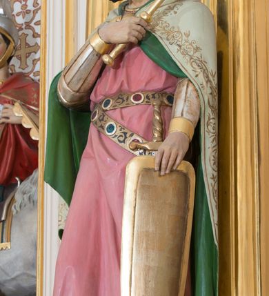 Zdjęcie nr 3: Rzeźba drążona od tyłu, ustawiona na prostopadłościennym postumencie. Święty Władysław ukazany frontalnie, w pozycji stojącej, w lekkim kontrapoście z ciężarem ciała przeniesionym na lewą nogę. Przedstawiony jako młody mężczyzna z twarzą okoloną bujnymi, ciemnobrązowymi włosami oraz krótką, gęstą brodą. Ubrany jest w strój rycerski z nagolennikami, długą różową tuniką przepasaną metalowym pasem, u którego podwieszony miecz oraz biały płaszcz z zieloną podszewką i złoconą, zamkniętą koronę królów węgierskich. Swoją lewą rękę wspiera na tarczy, w prawej ugiętej w łokciu i złożonej na piersi trzyma topór. Polichromia w odsłoniętych partiach ciała naturalistyczna, detale złocone.