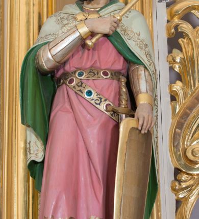 Zdjęcie nr 6: Rzeźba drążona od tyłu, ustawiona na prostopadłościennym postumencie. Święty Władysław ukazany frontalnie, w pozycji stojącej, w lekkim kontrapoście z ciężarem ciała przeniesionym na lewą nogę. Przedstawiony jako młody mężczyzna z twarzą okoloną bujnymi, ciemnobrązowymi włosami oraz krótką, gęstą brodą. Ubrany jest w strój rycerski z nagolennikami, długą różową tuniką przepasaną metalowym pasem, u którego podwieszony miecz oraz biały płaszcz z zieloną podszewką i złoconą, zamkniętą koronę królów węgierskich. Swoją lewą rękę wspiera na tarczy, w prawej ugiętej w łokciu i złożonej na piersi trzyma topór. Polichromia w odsłoniętych partiach ciała naturalistyczna, detale złocone.