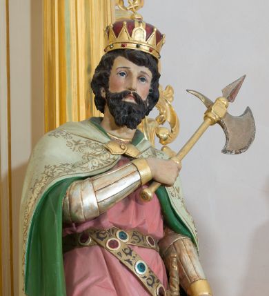 Zdjęcie nr 1: Rzeźba drążona od tyłu, ustawiona na prostopadłościennym postumencie. Święty Władysław ukazany frontalnie, w pozycji stojącej, w lekkim kontrapoście z ciężarem ciała przeniesionym na lewą nogę. Przedstawiony jako młody mężczyzna z twarzą okoloną bujnymi, ciemnobrązowymi włosami oraz krótką, gęstą brodą. Ubrany jest w strój rycerski z nagolennikami, długą różową tuniką przepasaną metalowym pasem, u którego podwieszony miecz oraz biały płaszcz z zieloną podszewką i złoconą, zamkniętą koronę królów węgierskich. Swoją lewą rękę wspiera na tarczy, w prawej ugiętej w łokciu i złożonej na piersi trzyma topór. Polichromia w odsłoniętych partiach ciała naturalistyczna, detale złocone.