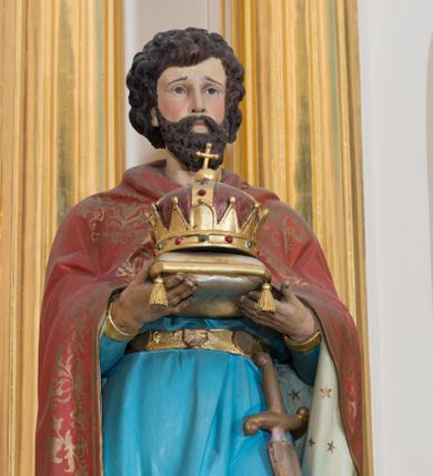 Zdjęcie nr 1: Rzeźba drążona od tyłu ustawiona na niskim, brązowym, prostopadłościennym postumencie. Święty ukazany jest frontalnie w pozycji stojącej, w lekkim kontrapoście z lewą nogą ugiętą w kolanie. Oburącz trzyma przed sobą złoconą koronę w typie zamkniętym ułożoną na poduszce. Przedstawiony jest jako młody mężczyzna z twarzą okoloną bujnymi, ciemnobrązowymi włosami oraz krótką i gęstą brodą. Twarz podłużna z długim nosem i dużymi oczami. Ubrany jest w strój rycerski z nagolennikami; krótką niebieską tunikę, przepasaną metalowym pasem, u którego podwieszony jest miecz oraz ciemnoczerwony płaszcz z białą podszewką zdobioną gwiazdami. Płaszcz na brzegu dekorowany jest szeroką stylizowaną wicią roślinną. Polichromia w odsłoniętych partiach ciała naturalistyczna, detale złocone.