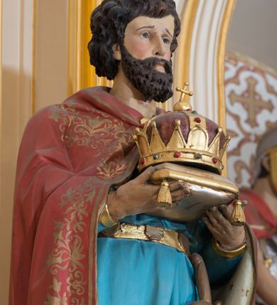 Zdjęcie nr 4: Rzeźba drążona od tyłu ustawiona na niskim, brązowym, prostopadłościennym postumencie. Święty ukazany jest frontalnie w pozycji stojącej, w lekkim kontrapoście z lewą nogą ugiętą w kolanie. Oburącz trzyma przed sobą złoconą koronę w typie zamkniętym ułożoną na poduszce. Przedstawiony jest jako młody mężczyzna z twarzą okoloną bujnymi, ciemnobrązowymi włosami oraz krótką i gęstą brodą. Twarz podłużna z długim nosem i dużymi oczami. Ubrany jest w strój rycerski z nagolennikami; krótką niebieską tunikę, przepasaną metalowym pasem, u którego podwieszony jest miecz oraz ciemnoczerwony płaszcz z białą podszewką zdobioną gwiazdami. Płaszcz na brzegu dekorowany jest szeroką stylizowaną wicią roślinną. Polichromia w odsłoniętych partiach ciała naturalistyczna, detale złocone.