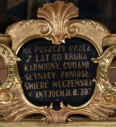 Zdjęcie nr 10: Ołtarz prostopadłościenny, górą wybrzuszony, z ukośnie dostawionymi cokołami, na których kanelowane wolnostojące filary z kompozytowymi kapitelami podtrzymujące wyłamane belkowanie z przerwanym przyczółkiem. Zwieńczenie z lekko wklęsłymi bokami ze spływami wolutowymi zakończone stykającymi się ze sobą wolutami. W partii belkowania kartusz ujęty karbowanymi wstęgami regencyjnymi, wolutami z listkami akantu i zwieńczony muszlą palmetową, w nim napis białą farbą na czarnym tle: „NA PUSZCZY PRZEZ / 7 LAT OD KRUKA / KARMIONY. CUDAMI / SŁYNĄCY. PONIÓSŁ / ŚMIERĆ MĘCZEŃSKĄ / W ANTJOCHJI R. 303”.