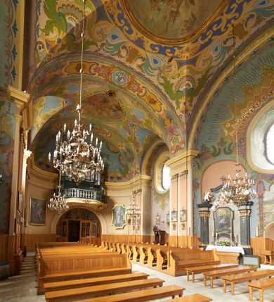 Zdjęcie nr 16: Polichromia pokrywa zarówno powierzchnię ścian, jak i sklepień kościoła, jednak zdecydowanie bardziej dekoracyjne motywy pojawiają się w górnych partiach murów. Dominuje stylizowana ornamentyka roślinna na błękitnym tle, utrzymana w jasnych, żywych tonacjach zróżnicowanych kolorów. Szerokie bordiury o tworzących je fantazyjnych formach i roślinności ujmują niewielkich rozmiarów sceny figuralne umiejscowione pośrodku sklepienia każdego z przęseł kościoła. Kompozycje, o zbliżonych do owalu kształtach przedstawiają: Śmierć św. Józefa w ostatnim przęśle nawy, Ukoronowanie Matki Boskiej na przecięciu nawy z transeptem oraz Wniebowzięcie Najświętszej Marii Panny w przęśle prezbiterium. W konsze absydy ukazana jest scena adoracji kielicha eucharystycznego przez anioły o tęczowych skrzydłach, ubrane w barwne, wzorzyste szaty. Po lewej stronie kościoła, na ścianie transeptu jest ukazana na tle pejzażu scena Chrztu Jezusa. 