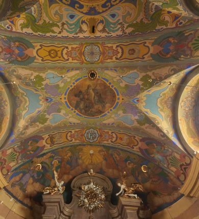 Zdjęcie nr 28: Polichromia pokrywa zarówno powierzchnię ścian, jak i sklepień kościoła, jednak zdecydowanie bardziej dekoracyjne motywy pojawiają się w górnych partiach murów. Dominuje stylizowana ornamentyka roślinna na błękitnym tle, utrzymana w jasnych, żywych tonacjach zróżnicowanych kolorów. Szerokie bordiury o tworzących je fantazyjnych formach i roślinności ujmują niewielkich rozmiarów sceny figuralne umiejscowione pośrodku sklepienia każdego z przęseł kościoła. Kompozycje, o zbliżonych do owalu kształtach przedstawiają: Śmierć św. Józefa w ostatnim przęśle nawy, Ukoronowanie Matki Boskiej na przecięciu nawy z transeptem oraz Wniebowzięcie Najświętszej Marii Panny w przęśle prezbiterium. W konsze absydy ukazana jest scena adoracji kielicha eucharystycznego przez anioły o tęczowych skrzydłach, ubrane w barwne, wzorzyste szaty. Po lewej stronie kościoła, na ścianie transeptu jest ukazana na tle pejzażu scena Chrztu Jezusa. 