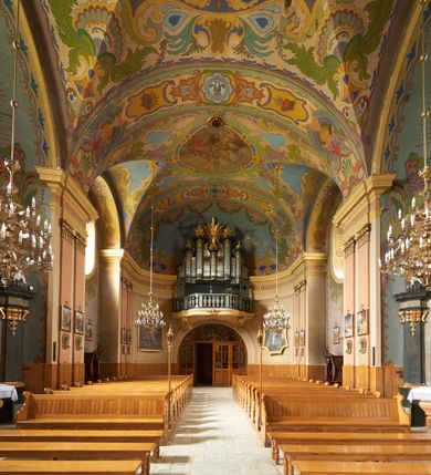 Zdjęcie nr 14: Polichromia pokrywa zarówno powierzchnię ścian, jak i sklepień kościoła, jednak zdecydowanie bardziej dekoracyjne motywy pojawiają się w górnych partiach murów. Dominuje stylizowana ornamentyka roślinna na błękitnym tle, utrzymana w jasnych, żywych tonacjach zróżnicowanych kolorów. Szerokie bordiury o tworzących je fantazyjnych formach i roślinności ujmują niewielkich rozmiarów sceny figuralne umiejscowione pośrodku sklepienia każdego z przęseł kościoła. Kompozycje, o zbliżonych do owalu kształtach przedstawiają: Śmierć św. Józefa w ostatnim przęśle nawy, Ukoronowanie Matki Boskiej na przecięciu nawy z transeptem oraz Wniebowzięcie Najświętszej Marii Panny w przęśle prezbiterium. W konsze absydy ukazana jest scena adoracji kielicha eucharystycznego przez anioły o tęczowych skrzydłach, ubrane w barwne, wzorzyste szaty. Po lewej stronie kościoła, na ścianie transeptu jest ukazana na tle pejzażu scena Chrztu Jezusa. 