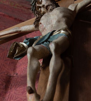Zdjęcie nr 9: Rzeźba ścięta z tyłu, przybita do krzyża trzema gwoździami, w typie Christo vivo. Ciało w lekkim zwisie o horyzontalnie rozłożonych ramionach, głowa opada na prawy bark, nogi lekko ugięte, stopy skrzyżowane w układzie prawa na lewą. Ciało jest silnie wysmuklone, z wyraźnie rozbudowaną i wypchniętą do przodu klatką piersiową, z podkreślonymi żebrami i zapadłym brzuchem; ręce i nogi są bardzo wychudzone z wyłamanymi barkami, podkreślonymi łokciami i kolanami. Twarz jest pociągła, wychudzona, z głęboko osadzonymi i rzeźbionymi, półotwartymi oczami, wąskim, długim nosem oraz otwartymi ustami z opadającą żuchwą; okolona krótką brodą, głęboko rzeźbioną w regularne pukle oraz długimi włosami ułożonymi w pojedyncze lekko skręcone pukle opadające na plecy oraz na klatkę piersiową. Głowę wieńczy masywna korona cierniowa. Perizonium jest krótkie, silnie opasujące ciało, zawiązane w partii podbrzusza z rozwianym ekspresyjnie fragmentem tkaniny w charakterystycznym odwinięciu na prawym boku. Figura jest polichromowana naturalistycznie, ciało wyróżnia się bladą karnacją z zaznaczonymi śladami męki; perizonium jest złocone i niebieskie na podszewce.