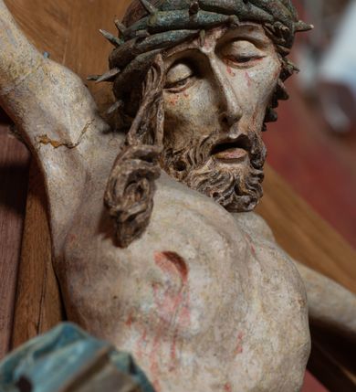 Zdjęcie nr 14: Rzeźba ścięta z tyłu, przybita do krzyża trzema gwoździami, w typie Christo vivo. Ciało w lekkim zwisie o horyzontalnie rozłożonych ramionach, głowa opada na prawy bark, nogi lekko ugięte, stopy skrzyżowane w układzie prawa na lewą. Ciało jest silnie wysmuklone, z wyraźnie rozbudowaną i wypchniętą do przodu klatką piersiową, z podkreślonymi żebrami i zapadłym brzuchem; ręce i nogi są bardzo wychudzone z wyłamanymi barkami, podkreślonymi łokciami i kolanami. Twarz jest pociągła, wychudzona, z głęboko osadzonymi i rzeźbionymi, półotwartymi oczami, wąskim, długim nosem oraz otwartymi ustami z opadającą żuchwą; okolona krótką brodą, głęboko rzeźbioną w regularne pukle oraz długimi włosami ułożonymi w pojedyncze lekko skręcone pukle opadające na plecy oraz na klatkę piersiową. Głowę wieńczy masywna korona cierniowa. Perizonium jest krótkie, silnie opasujące ciało, zawiązane w partii podbrzusza z rozwianym ekspresyjnie fragmentem tkaniny w charakterystycznym odwinięciu na prawym boku. Figura jest polichromowana naturalistycznie, ciało wyróżnia się bladą karnacją z zaznaczonymi śladami męki; perizonium jest złocone i niebieskie na podszewce.