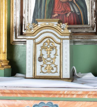 Zdjęcie nr 1: Tabernakulum ma formę prostopadłościennej szafki na niskim cokoliku, zwieńczonej belkowaniem w formie półkolistego łuku nadwieszonego. Drzwiczki mają kształt stojącego prostokąta zamkniętego łukiem wklęsło-wypukłym, po bokach ujęte są parą listewek. Na tabernakulum nałożony niewysoki cokolik pełniący funkcję tronu wystawienia. Pole drzwiczek zdobi stylizowany motyw roślinny o symetrycznym układzie. Górna krawędź gzymsu wieńczącego zdobiona powtarzającym się motywem trójliścia oraz parą gałązek różanych. Struktura tabernakulum jest srebrzona, ornamentyka i detal architektoniczny złocone.      