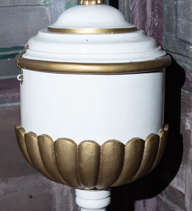 Zdjęcie nr 1: Wolnostojąca chrzcielnica w typie kielichowym osadzona jest na okrągłej, profilowanej, wklęsło-wypukłej stopie. Toczony trzon w dolnej partii zdobi gruszkowy nodus przechodzący w ujęty dwoma pierścieniami (talerzykami) wałek. Czara jest cylindryczna, dołem puklowana, o profilowanym wylewie. Struktura czaszy chrzcielnicy jest profilowana, wklęsło-wypukła i zwieńczona złoconym krzyżem o trójlistnych zakończeniach ramion. Całość utrzymana jest w kolorze bieli, część detali jest złocona. 