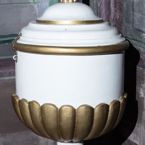 Zdjęcie nr 1: Wolnostojąca chrzcielnica w typie kielichowym osadzona jest na okrągłej, profilowanej, wklęsło-wypukłej stopie. Toczony trzon w dolnej partii zdobi gruszkowy nodus przechodzący w ujęty dwoma pierścieniami (talerzykami) wałek. Czara jest cylindryczna, dołem puklowana, o profilowanym wylewie. Struktura czaszy chrzcielnicy jest profilowana, wklęsło-wypukła i zwieńczona złoconym krzyżem o trójlistnych zakończeniach ramion. Całość utrzymana jest w kolorze bieli, część detali jest złocona. 