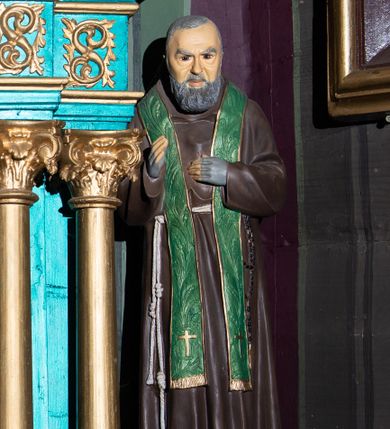 Zdjęcie nr 1: Pełnoplastyczna figura św. Ojca Pio ukazanego w całej postaci, stojącego frontalnie, na niewysokiej podstawie zbliżonej do prostopadłościanu. Głowa jest lekko pochylona w dół. Prawa ręka uniesiona jest w geście błogosławieństwa, lewa wsparta na piersiach. Pociągłą twarz charakteryzują rysy dojrzałego mężczyzny, ciemne, podkrążone oczy oraz półdługi, gęsty, siwy zarost; na głowie są krótkie, siwe włosy. Święty ubrany jest w habit kapucyński przewiązany sznurem, na którym zawieszony jest czarny różaniec. Na ramiona nałożona jest zielona stuła zdobiona reliefowym motywem kłosów zbóż. Dłonie osłaniają szare rękawiczki w typie mitenek. Na stopach są ciemnobrązowe buty z kwadratowymi noskami. Na podstawie znajduje się niewielka tabliczka w formie banderoli z napisem: „O(jciec) PIO”. 