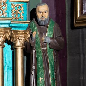 Zdjęcie nr 1: Pełnoplastyczna figura św. Ojca Pio ukazanego w całej postaci, stojącego frontalnie, na niewysokiej podstawie zbliżonej do prostopadłościanu. Głowa jest lekko pochylona w dół. Prawa ręka uniesiona jest w geście błogosławieństwa, lewa wsparta na piersiach. Pociągłą twarz charakteryzują rysy dojrzałego mężczyzny, ciemne, podkrążone oczy oraz półdługi, gęsty, siwy zarost; na głowie są krótkie, siwe włosy. Święty ubrany jest w habit kapucyński przewiązany sznurem, na którym zawieszony jest czarny różaniec. Na ramiona nałożona jest zielona stuła zdobiona reliefowym motywem kłosów zbóż. Dłonie osłaniają szare rękawiczki w typie mitenek. Na stopach są ciemnobrązowe buty z kwadratowymi noskami. Na podstawie znajduje się niewielka tabliczka w formie banderoli z napisem: „O(jciec) PIO”. 