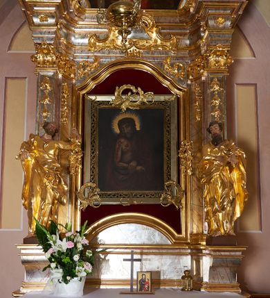 Zdjęcie nr 1: Nastawa ołtarzowa architektoniczna, jednoosiowa, jednokondygnacyjna ze zwieńczeniem. Mensa drewniana o profilowanej krawędzi. Ołtarz  sarkofagowy, na profilowanym cokole; na froncie intarsjowany wzór geometryczny. Kondygnacja ustawiona na profilowanym cokole, ujęta parą zdwojonych filarów o kanelowanych trzonach i kapitelach kompozytowych. Podpory dźwigają pełne belkowanie, wyłamane na ich osi, o wklęsło-wypukłym fryzie i silnie wysuniętym gzymsie oraz przerwany wolutowy przyczółek. W polu środkowym profilowana rama w formie stojącego prostokąta zamkniętego łukiem segmentowym nadwieszonym z uskokiem, z mniejszym obrazem wewnątrz w formie stojącego prostokąta, z przedstawieniem Ecce Homo. Przed filarami, na wklęsło-wypukłych postumentach dwie figury niezidentyfikowanych świętych. Zwieńczenie w formie leżącego prostokąta ujętego spływami wolutowymi, zamkniętego falistym gzymsem przechodzącym w zwrócone przeciwstawnie woluty. W polu rama o fantazyjnej formie zbliżonej do trójkąta z obrazem przedstawiającym Narodzenie Marii. Trzony filarów dekorowane kampanulami. W polu środkowym w przyłuczach rocaille. Rama obrazu na dłuższych bokach zdobiona ornamentem rocaille'owym. Nad polem środkowym w polu belkowania racaille'owy kartusz  z napisem: „ECCE HOMO”. W partii fryzu, na osiach podpór z trzech stron rozety. Struktura w całości laserunkowa, srebrzona, marmoryzowana; ornamentyka i profile złocone.