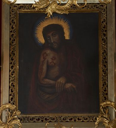 Zdjęcie nr 1: Obraz w kształcie stojącego prostokąta ujęty profilowaną, srebrzoną ramą, od wewnątrz zdobioną ażurowym ornamentem. Przedstawienie Chrystusa w półpostaci, zwróconego w trzech czwartych w prawo  z dłońmi skrzyżowanymi na wysokości pasa. Twarz podłużna o wyrazistych, pociągłych rysach z krótkim, brązowym zarostem. Włosy długie, falowane, opadające na plecy i ramiona. Kolorystyka ciała ciemna. Na głowie cierniowa korona. Na czole, szyi, ramionach i klatce piersiowej obfite krople krwi. Biodra przewiązane białym perizonium, plecy osłonięte ciemnoczerwonym płaszczem, przewieszonym przez lewą rękę, odsłaniającym nagi tors. Tło ciemnobrązowe, wokół głowy złoty promienisty nimb i jasnoniebieski otok. 