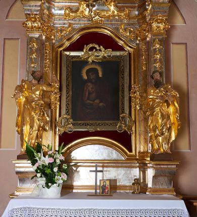 Zdjęcie nr 2: Obraz w kształcie stojącego prostokąta ujęty profilowaną, srebrzoną ramą, od wewnątrz zdobioną ażurowym ornamentem. Przedstawienie Chrystusa w półpostaci, zwróconego w trzech czwartych w prawo  z dłońmi skrzyżowanymi na wysokości pasa. Twarz podłużna o wyrazistych, pociągłych rysach z krótkim, brązowym zarostem. Włosy długie, falowane, opadające na plecy i ramiona. Kolorystyka ciała ciemna. Na głowie cierniowa korona. Na czole, szyi, ramionach i klatce piersiowej obfite krople krwi. Biodra przewiązane białym perizonium, plecy osłonięte ciemnoczerwonym płaszczem, przewieszonym przez lewą rękę, odsłaniającym nagi tors. Tło ciemnobrązowe, wokół głowy złoty promienisty nimb i jasnoniebieski otok. 