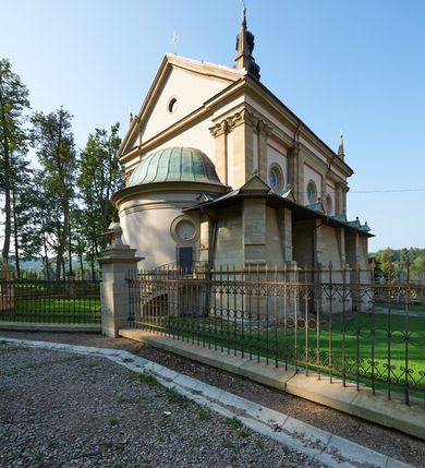 Zdjęcie nr 24: Kościół należy do zespołu dróżek Matki Boskiej, położnych w obrębie klasztoru w Kalwarii Zebrzydowskiej. Świątynia jest położna w centrum wsi, na wschód od głównej drogi, na placu otoczonym kamiennym murem z żeliwnym płotem, który od frontu ozdobiony jest figurami 12 apostołów, umieszczonymi na wysokich cokołach. Główne wejście na plac prowadzi przez bramę od zachodu, flankowaną dwoma filarami, na których ustawione są klasycystyczne wazony. Drugie wejście usytuowane jest od wschodu przez murowaną bramę, nakrytą daszkiem dwuspadowym, na którym ustawiona jest figura grającego na trąbie anioła. Trzecie wejście znajduje się od północy.

Kościół jest budowlą orientowaną, założoną na planie prostokąta, jednonawową, zamkniętą półkolistą absydą. Od zachodu przylega kruchta na planie prostokąta, zamkniętego z jednej strony półkoliście. Wewnątrz kościół dwupiętrowy. Na parterze część wschodnia świątyni podniesiona jest o siedem stopni, oddzielona od nawy trzema półkolistymi arkadami, wspartymi na czterech filarach i ustawionymi przed nimi półkolumnami, które wspierają pełne belkowanie, wyłamane na osiach. W przyłuczach płskorzeźbione główki aniołków. Na fryzie złocony napis: „EIA ADVOCATA NOSTRA OCVLOS MISERICORDES AD NOS CONVERTE ET IESVM BENEDICTV FRCTVM NOBIS OSTENDE”. Za środkową arkadą usytuowana jest niewielka, najstarsza część świątyni – Grób Matki Boskiej, na rzucie prostokąta, sklepiony krzyżowo. W ścianach bocznych nawy są półkoliście zamknięte wnęki na ołtarze. Od zachodu chór muzyczny wsparty jest na trzech arkadach filarowych. Wnętrze obiega drewniany ganek wsparty na metalowych wspornikach, a w części wschodniej i zachodniej na trzech murowanych arkadach. Podniebienie ganku zdobione jest geometrycznym stiukiem. W części nawowej świątyni znajduje się sklepienie kolebkowe z lunetami, w apsydzie hemisferyczne. Wnętrze doświetlone jest sześcioma małymi, okrągłymi oknami znajdującymi się na piętrze od strony północnej i południowej.

Fasada w dolnej części zaakcentowana jest przez monumentalną kruchtę w formie aediculi, założoną na rzucie prostokąta zamkniętego półkoliście. Ściany artykułowane są pięcioma półkolistymi arkadami, wspartymi na filarkach. Od zewnątrz osie ujęte są przez kanelowane kolumny, ustawione na wysokich cokołach, na tle pilastrów. Podpory wznoszą pełne belkowanie i wysunięty gzyms zdobiony kimationem i dekoracją kostkową. Nakrycie w formie kopuły półkolistej z latarnią. Belkowanie ozdobione jest dekoracją roślinną i główkami aniołków. Po bokach kruchty wysunięte są fragmenty muru oporowego do połowy wysokości fasady. Powyżej fasada flankowana przez dwie pary pilastrów z jońskimi kapitelami wspierającymi niepełne belkowanie. Pośrodku duże, półkoliste okno z metalową, dekoracyjną kratą. Szczyt trójkątny z oculusem pośrodku. Na szczycie ustawiona jest figura Najświętszej Marii Panny. Świątynia dookoła obwiedziona szerokim murem oporowym, nakrytym daszkiem, o ukośnych ścianach, flankowanych szkarpami, po sześć na każdej ścianie bocznej i dwoma na ścianie zachodniej oraz wschodniej. Powyżej elewacje ujęte pilastrami z kapitelami jońskimi wznoszącymi niepełne belkowanie. Na wschodniej ścianie półkolista apsyda, nakryta półkopułą. Kościół nakryty dachem siodłowym ze zdwojoną, wysoką latarnią pośrodku. Na narożach dachu ustawione wysokie obeliski na cokołach. 

 