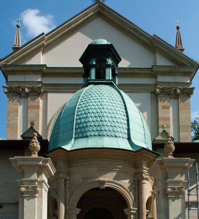 Zdjęcie nr 9: Kościół należy do zespołu dróżek Matki Boskiej, położnych w obrębie klasztoru w Kalwarii Zebrzydowskiej. Świątynia jest położna w centrum wsi, na wschód od głównej drogi, na placu otoczonym kamiennym murem z żeliwnym płotem, który od frontu ozdobiony jest figurami 12 apostołów, umieszczonymi na wysokich cokołach. Główne wejście na plac prowadzi przez bramę od zachodu, flankowaną dwoma filarami, na których ustawione są klasycystyczne wazony. Drugie wejście usytuowane jest od wschodu przez murowaną bramę, nakrytą daszkiem dwuspadowym, na którym ustawiona jest figura grającego na trąbie anioła. Trzecie wejście znajduje się od północy.

Kościół jest budowlą orientowaną, założoną na planie prostokąta, jednonawową, zamkniętą półkolistą absydą. Od zachodu przylega kruchta na planie prostokąta, zamkniętego z jednej strony półkoliście. Wewnątrz kościół dwupiętrowy. Na parterze część wschodnia świątyni podniesiona jest o siedem stopni, oddzielona od nawy trzema półkolistymi arkadami, wspartymi na czterech filarach i ustawionymi przed nimi półkolumnami, które wspierają pełne belkowanie, wyłamane na osiach. W przyłuczach płskorzeźbione główki aniołków. Na fryzie złocony napis: „EIA ADVOCATA NOSTRA OCVLOS MISERICORDES AD NOS CONVERTE ET IESVM BENEDICTV FRCTVM NOBIS OSTENDE”. Za środkową arkadą usytuowana jest niewielka, najstarsza część świątyni – Grób Matki Boskiej, na rzucie prostokąta, sklepiony krzyżowo. W ścianach bocznych nawy są półkoliście zamknięte wnęki na ołtarze. Od zachodu chór muzyczny wsparty jest na trzech arkadach filarowych. Wnętrze obiega drewniany ganek wsparty na metalowych wspornikach, a w części wschodniej i zachodniej na trzech murowanych arkadach. Podniebienie ganku zdobione jest geometrycznym stiukiem. W części nawowej świątyni znajduje się sklepienie kolebkowe z lunetami, w apsydzie hemisferyczne. Wnętrze doświetlone jest sześcioma małymi, okrągłymi oknami znajdującymi się na piętrze od strony północnej i południowej.

Fasada w dolnej części zaakcentowana jest przez monumentalną kruchtę w formie aediculi, założoną na rzucie prostokąta zamkniętego półkoliście. Ściany artykułowane są pięcioma półkolistymi arkadami, wspartymi na filarkach. Od zewnątrz osie ujęte są przez kanelowane kolumny, ustawione na wysokich cokołach, na tle pilastrów. Podpory wznoszą pełne belkowanie i wysunięty gzyms zdobiony kimationem i dekoracją kostkową. Nakrycie w formie kopuły półkolistej z latarnią. Belkowanie ozdobione jest dekoracją roślinną i główkami aniołków. Po bokach kruchty wysunięte są fragmenty muru oporowego do połowy wysokości fasady. Powyżej fasada flankowana przez dwie pary pilastrów z jońskimi kapitelami wspierającymi niepełne belkowanie. Pośrodku duże, półkoliste okno z metalową, dekoracyjną kratą. Szczyt trójkątny z oculusem pośrodku. Na szczycie ustawiona jest figura Najświętszej Marii Panny. Świątynia dookoła obwiedziona szerokim murem oporowym, nakrytym daszkiem, o ukośnych ścianach, flankowanych szkarpami, po sześć na każdej ścianie bocznej i dwoma na ścianie zachodniej oraz wschodniej. Powyżej elewacje ujęte pilastrami z kapitelami jońskimi wznoszącymi niepełne belkowanie. Na wschodniej ścianie półkolista apsyda, nakryta półkopułą. Kościół nakryty dachem siodłowym ze zdwojoną, wysoką latarnią pośrodku. Na narożach dachu ustawione wysokie obeliski na cokołach. 

 