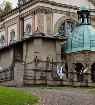 Zdjęcie nr 5: Kościół należy do zespołu dróżek Matki Boskiej, położnych w obrębie klasztoru w Kalwarii Zebrzydowskiej. Świątynia jest położna w centrum wsi, na wschód od głównej drogi, na placu otoczonym kamiennym murem z żeliwnym płotem, który od frontu ozdobiony jest figurami 12 apostołów, umieszczonymi na wysokich cokołach. Główne wejście na plac prowadzi przez bramę od zachodu, flankowaną dwoma filarami, na których ustawione są klasycystyczne wazony. Drugie wejście usytuowane jest od wschodu przez murowaną bramę, nakrytą daszkiem dwuspadowym, na którym ustawiona jest figura grającego na trąbie anioła. Trzecie wejście znajduje się od północy.

Kościół jest budowlą orientowaną, założoną na planie prostokąta, jednonawową, zamkniętą półkolistą absydą. Od zachodu przylega kruchta na planie prostokąta, zamkniętego z jednej strony półkoliście. Wewnątrz kościół dwupiętrowy. Na parterze część wschodnia świątyni podniesiona jest o siedem stopni, oddzielona od nawy trzema półkolistymi arkadami, wspartymi na czterech filarach i ustawionymi przed nimi półkolumnami, które wspierają pełne belkowanie, wyłamane na osiach. W przyłuczach płskorzeźbione główki aniołków. Na fryzie złocony napis: „EIA ADVOCATA NOSTRA OCVLOS MISERICORDES AD NOS CONVERTE ET IESVM BENEDICTV FRCTVM NOBIS OSTENDE”. Za środkową arkadą usytuowana jest niewielka, najstarsza część świątyni – Grób Matki Boskiej, na rzucie prostokąta, sklepiony krzyżowo. W ścianach bocznych nawy są półkoliście zamknięte wnęki na ołtarze. Od zachodu chór muzyczny wsparty jest na trzech arkadach filarowych. Wnętrze obiega drewniany ganek wsparty na metalowych wspornikach, a w części wschodniej i zachodniej na trzech murowanych arkadach. Podniebienie ganku zdobione jest geometrycznym stiukiem. W części nawowej świątyni znajduje się sklepienie kolebkowe z lunetami, w apsydzie hemisferyczne. Wnętrze doświetlone jest sześcioma małymi, okrągłymi oknami znajdującymi się na piętrze od strony północnej i południowej.

Fasada w dolnej części zaakcentowana jest przez monumentalną kruchtę w formie aediculi, założoną na rzucie prostokąta zamkniętego półkoliście. Ściany artykułowane są pięcioma półkolistymi arkadami, wspartymi na filarkach. Od zewnątrz osie ujęte są przez kanelowane kolumny, ustawione na wysokich cokołach, na tle pilastrów. Podpory wznoszą pełne belkowanie i wysunięty gzyms zdobiony kimationem i dekoracją kostkową. Nakrycie w formie kopuły półkolistej z latarnią. Belkowanie ozdobione jest dekoracją roślinną i główkami aniołków. Po bokach kruchty wysunięte są fragmenty muru oporowego do połowy wysokości fasady. Powyżej fasada flankowana przez dwie pary pilastrów z jońskimi kapitelami wspierającymi niepełne belkowanie. Pośrodku duże, półkoliste okno z metalową, dekoracyjną kratą. Szczyt trójkątny z oculusem pośrodku. Na szczycie ustawiona jest figura Najświętszej Marii Panny. Świątynia dookoła obwiedziona szerokim murem oporowym, nakrytym daszkiem, o ukośnych ścianach, flankowanych szkarpami, po sześć na każdej ścianie bocznej i dwoma na ścianie zachodniej oraz wschodniej. Powyżej elewacje ujęte pilastrami z kapitelami jońskimi wznoszącymi niepełne belkowanie. Na wschodniej ścianie półkolista apsyda, nakryta półkopułą. Kościół nakryty dachem siodłowym ze zdwojoną, wysoką latarnią pośrodku. Na narożach dachu ustawione wysokie obeliski na cokołach. 

 