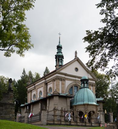 Zdjęcie nr 68: Kościół należy do zespołu dróżek Matki Boskiej, położnych w obrębie klasztoru w Kalwarii Zebrzydowskiej. Świątynia jest położna w centrum wsi, na wschód od głównej drogi, na placu otoczonym kamiennym murem z żeliwnym płotem, który od frontu ozdobiony jest figurami 12 apostołów, umieszczonymi na wysokich cokołach. Główne wejście na plac prowadzi przez bramę od zachodu, flankowaną dwoma filarami, na których ustawione są klasycystyczne wazony. Drugie wejście usytuowane jest od wschodu przez murowaną bramę, nakrytą daszkiem dwuspadowym, na którym ustawiona jest figura grającego na trąbie anioła. Trzecie wejście znajduje się od północy.

Kościół jest budowlą orientowaną, założoną na planie prostokąta, jednonawową, zamkniętą półkolistą absydą. Od zachodu przylega kruchta na planie prostokąta, zamkniętego z jednej strony półkoliście. Wewnątrz kościół dwupiętrowy. Na parterze część wschodnia świątyni podniesiona jest o siedem stopni, oddzielona od nawy trzema półkolistymi arkadami, wspartymi na czterech filarach i ustawionymi przed nimi półkolumnami, które wspierają pełne belkowanie, wyłamane na osiach. W przyłuczach płskorzeźbione główki aniołków. Na fryzie złocony napis: „EIA ADVOCATA NOSTRA OCVLOS MISERICORDES AD NOS CONVERTE ET IESVM BENEDICTV FRCTVM NOBIS OSTENDE”. Za środkową arkadą usytuowana jest niewielka, najstarsza część świątyni – Grób Matki Boskiej, na rzucie prostokąta, sklepiony krzyżowo. W ścianach bocznych nawy są półkoliście zamknięte wnęki na ołtarze. Od zachodu chór muzyczny wsparty jest na trzech arkadach filarowych. Wnętrze obiega drewniany ganek wsparty na metalowych wspornikach, a w części wschodniej i zachodniej na trzech murowanych arkadach. Podniebienie ganku zdobione jest geometrycznym stiukiem. W części nawowej świątyni znajduje się sklepienie kolebkowe z lunetami, w apsydzie hemisferyczne. Wnętrze doświetlone jest sześcioma małymi, okrągłymi oknami znajdującymi się na piętrze od strony północnej i południowej.

Fasada w dolnej części zaakcentowana jest przez monumentalną kruchtę w formie aediculi, założoną na rzucie prostokąta zamkniętego półkoliście. Ściany artykułowane są pięcioma półkolistymi arkadami, wspartymi na filarkach. Od zewnątrz osie ujęte są przez kanelowane kolumny, ustawione na wysokich cokołach, na tle pilastrów. Podpory wznoszą pełne belkowanie i wysunięty gzyms zdobiony kimationem i dekoracją kostkową. Nakrycie w formie kopuły półkolistej z latarnią. Belkowanie ozdobione jest dekoracją roślinną i główkami aniołków. Po bokach kruchty wysunięte są fragmenty muru oporowego do połowy wysokości fasady. Powyżej fasada flankowana przez dwie pary pilastrów z jońskimi kapitelami wspierającymi niepełne belkowanie. Pośrodku duże, półkoliste okno z metalową, dekoracyjną kratą. Szczyt trójkątny z oculusem pośrodku. Na szczycie ustawiona jest figura Najświętszej Marii Panny. Świątynia dookoła obwiedziona szerokim murem oporowym, nakrytym daszkiem, o ukośnych ścianach, flankowanych szkarpami, po sześć na każdej ścianie bocznej i dwoma na ścianie zachodniej oraz wschodniej. Powyżej elewacje ujęte pilastrami z kapitelami jońskimi wznoszącymi niepełne belkowanie. Na wschodniej ścianie półkolista apsyda, nakryta półkopułą. Kościół nakryty dachem siodłowym ze zdwojoną, wysoką latarnią pośrodku. Na narożach dachu ustawione wysokie obeliski na cokołach. 

 