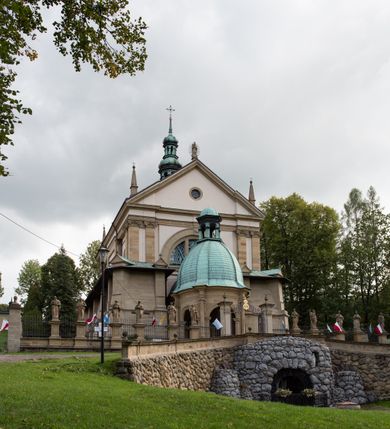 Zdjęcie nr 46: Kościół należy do zespołu dróżek Matki Boskiej, położnych w obrębie klasztoru w Kalwarii Zebrzydowskiej. Świątynia jest położna w centrum wsi, na wschód od głównej drogi, na placu otoczonym kamiennym murem z żeliwnym płotem, który od frontu ozdobiony jest figurami 12 apostołów, umieszczonymi na wysokich cokołach. Główne wejście na plac prowadzi przez bramę od zachodu, flankowaną dwoma filarami, na których ustawione są klasycystyczne wazony. Drugie wejście usytuowane jest od wschodu przez murowaną bramę, nakrytą daszkiem dwuspadowym, na którym ustawiona jest figura grającego na trąbie anioła. Trzecie wejście znajduje się od północy.

Kościół jest budowlą orientowaną, założoną na planie prostokąta, jednonawową, zamkniętą półkolistą absydą. Od zachodu przylega kruchta na planie prostokąta, zamkniętego z jednej strony półkoliście. Wewnątrz kościół dwupiętrowy. Na parterze część wschodnia świątyni podniesiona jest o siedem stopni, oddzielona od nawy trzema półkolistymi arkadami, wspartymi na czterech filarach i ustawionymi przed nimi półkolumnami, które wspierają pełne belkowanie, wyłamane na osiach. W przyłuczach płskorzeźbione główki aniołków. Na fryzie złocony napis: „EIA ADVOCATA NOSTRA OCVLOS MISERICORDES AD NOS CONVERTE ET IESVM BENEDICTV FRCTVM NOBIS OSTENDE”. Za środkową arkadą usytuowana jest niewielka, najstarsza część świątyni – Grób Matki Boskiej, na rzucie prostokąta, sklepiony krzyżowo. W ścianach bocznych nawy są półkoliście zamknięte wnęki na ołtarze. Od zachodu chór muzyczny wsparty jest na trzech arkadach filarowych. Wnętrze obiega drewniany ganek wsparty na metalowych wspornikach, a w części wschodniej i zachodniej na trzech murowanych arkadach. Podniebienie ganku zdobione jest geometrycznym stiukiem. W części nawowej świątyni znajduje się sklepienie kolebkowe z lunetami, w apsydzie hemisferyczne. Wnętrze doświetlone jest sześcioma małymi, okrągłymi oknami znajdującymi się na piętrze od strony północnej i południowej.

Fasada w dolnej części zaakcentowana jest przez monumentalną kruchtę w formie aediculi, założoną na rzucie prostokąta zamkniętego półkoliście. Ściany artykułowane są pięcioma półkolistymi arkadami, wspartymi na filarkach. Od zewnątrz osie ujęte są przez kanelowane kolumny, ustawione na wysokich cokołach, na tle pilastrów. Podpory wznoszą pełne belkowanie i wysunięty gzyms zdobiony kimationem i dekoracją kostkową. Nakrycie w formie kopuły półkolistej z latarnią. Belkowanie ozdobione jest dekoracją roślinną i główkami aniołków. Po bokach kruchty wysunięte są fragmenty muru oporowego do połowy wysokości fasady. Powyżej fasada flankowana przez dwie pary pilastrów z jońskimi kapitelami wspierającymi niepełne belkowanie. Pośrodku duże, półkoliste okno z metalową, dekoracyjną kratą. Szczyt trójkątny z oculusem pośrodku. Na szczycie ustawiona jest figura Najświętszej Marii Panny. Świątynia dookoła obwiedziona szerokim murem oporowym, nakrytym daszkiem, o ukośnych ścianach, flankowanych szkarpami, po sześć na każdej ścianie bocznej i dwoma na ścianie zachodniej oraz wschodniej. Powyżej elewacje ujęte pilastrami z kapitelami jońskimi wznoszącymi niepełne belkowanie. Na wschodniej ścianie półkolista apsyda, nakryta półkopułą. Kościół nakryty dachem siodłowym ze zdwojoną, wysoką latarnią pośrodku. Na narożach dachu ustawione wysokie obeliski na cokołach. 

 
