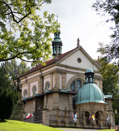 Zdjęcie nr 79: Kościół należy do zespołu dróżek Matki Boskiej, położnych w obrębie klasztoru w Kalwarii Zebrzydowskiej. Świątynia jest położna w centrum wsi, na wschód od głównej drogi, na placu otoczonym kamiennym murem z żeliwnym płotem, który od frontu ozdobiony jest figurami 12 apostołów, umieszczonymi na wysokich cokołach. Główne wejście na plac prowadzi przez bramę od zachodu, flankowaną dwoma filarami, na których ustawione są klasycystyczne wazony. Drugie wejście usytuowane jest od wschodu przez murowaną bramę, nakrytą daszkiem dwuspadowym, na którym ustawiona jest figura grającego na trąbie anioła. Trzecie wejście znajduje się od północy.

Kościół jest budowlą orientowaną, założoną na planie prostokąta, jednonawową, zamkniętą półkolistą absydą. Od zachodu przylega kruchta na planie prostokąta, zamkniętego z jednej strony półkoliście. Wewnątrz kościół dwupiętrowy. Na parterze część wschodnia świątyni podniesiona jest o siedem stopni, oddzielona od nawy trzema półkolistymi arkadami, wspartymi na czterech filarach i ustawionymi przed nimi półkolumnami, które wspierają pełne belkowanie, wyłamane na osiach. W przyłuczach płskorzeźbione główki aniołków. Na fryzie złocony napis: „EIA ADVOCATA NOSTRA OCVLOS MISERICORDES AD NOS CONVERTE ET IESVM BENEDICTV FRCTVM NOBIS OSTENDE”. Za środkową arkadą usytuowana jest niewielka, najstarsza część świątyni – Grób Matki Boskiej, na rzucie prostokąta, sklepiony krzyżowo. W ścianach bocznych nawy są półkoliście zamknięte wnęki na ołtarze. Od zachodu chór muzyczny wsparty jest na trzech arkadach filarowych. Wnętrze obiega drewniany ganek wsparty na metalowych wspornikach, a w części wschodniej i zachodniej na trzech murowanych arkadach. Podniebienie ganku zdobione jest geometrycznym stiukiem. W części nawowej świątyni znajduje się sklepienie kolebkowe z lunetami, w apsydzie hemisferyczne. Wnętrze doświetlone jest sześcioma małymi, okrągłymi oknami znajdującymi się na piętrze od strony północnej i południowej.

Fasada w dolnej części zaakcentowana jest przez monumentalną kruchtę w formie aediculi, założoną na rzucie prostokąta zamkniętego półkoliście. Ściany artykułowane są pięcioma półkolistymi arkadami, wspartymi na filarkach. Od zewnątrz osie ujęte są przez kanelowane kolumny, ustawione na wysokich cokołach, na tle pilastrów. Podpory wznoszą pełne belkowanie i wysunięty gzyms zdobiony kimationem i dekoracją kostkową. Nakrycie w formie kopuły półkolistej z latarnią. Belkowanie ozdobione jest dekoracją roślinną i główkami aniołków. Po bokach kruchty wysunięte są fragmenty muru oporowego do połowy wysokości fasady. Powyżej fasada flankowana przez dwie pary pilastrów z jońskimi kapitelami wspierającymi niepełne belkowanie. Pośrodku duże, półkoliste okno z metalową, dekoracyjną kratą. Szczyt trójkątny z oculusem pośrodku. Na szczycie ustawiona jest figura Najświętszej Marii Panny. Świątynia dookoła obwiedziona szerokim murem oporowym, nakrytym daszkiem, o ukośnych ścianach, flankowanych szkarpami, po sześć na każdej ścianie bocznej i dwoma na ścianie zachodniej oraz wschodniej. Powyżej elewacje ujęte pilastrami z kapitelami jońskimi wznoszącymi niepełne belkowanie. Na wschodniej ścianie półkolista apsyda, nakryta półkopułą. Kościół nakryty dachem siodłowym ze zdwojoną, wysoką latarnią pośrodku. Na narożach dachu ustawione wysokie obeliski na cokołach. 

 