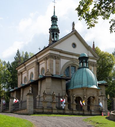 Zdjęcie nr 18: Kościół należy do zespołu dróżek Matki Boskiej, położnych w obrębie klasztoru w Kalwarii Zebrzydowskiej. Świątynia jest położna w centrum wsi, na wschód od głównej drogi, na placu otoczonym kamiennym murem z żeliwnym płotem, który od frontu ozdobiony jest figurami 12 apostołów, umieszczonymi na wysokich cokołach. Główne wejście na plac prowadzi przez bramę od zachodu, flankowaną dwoma filarami, na których ustawione są klasycystyczne wazony. Drugie wejście usytuowane jest od wschodu przez murowaną bramę, nakrytą daszkiem dwuspadowym, na którym ustawiona jest figura grającego na trąbie anioła. Trzecie wejście znajduje się od północy.

Kościół jest budowlą orientowaną, założoną na planie prostokąta, jednonawową, zamkniętą półkolistą absydą. Od zachodu przylega kruchta na planie prostokąta, zamkniętego z jednej strony półkoliście. Wewnątrz kościół dwupiętrowy. Na parterze część wschodnia świątyni podniesiona jest o siedem stopni, oddzielona od nawy trzema półkolistymi arkadami, wspartymi na czterech filarach i ustawionymi przed nimi półkolumnami, które wspierają pełne belkowanie, wyłamane na osiach. W przyłuczach płskorzeźbione główki aniołków. Na fryzie złocony napis: „EIA ADVOCATA NOSTRA OCVLOS MISERICORDES AD NOS CONVERTE ET IESVM BENEDICTV FRCTVM NOBIS OSTENDE”. Za środkową arkadą usytuowana jest niewielka, najstarsza część świątyni – Grób Matki Boskiej, na rzucie prostokąta, sklepiony krzyżowo. W ścianach bocznych nawy są półkoliście zamknięte wnęki na ołtarze. Od zachodu chór muzyczny wsparty jest na trzech arkadach filarowych. Wnętrze obiega drewniany ganek wsparty na metalowych wspornikach, a w części wschodniej i zachodniej na trzech murowanych arkadach. Podniebienie ganku zdobione jest geometrycznym stiukiem. W części nawowej świątyni znajduje się sklepienie kolebkowe z lunetami, w apsydzie hemisferyczne. Wnętrze doświetlone jest sześcioma małymi, okrągłymi oknami znajdującymi się na piętrze od strony północnej i południowej.

Fasada w dolnej części zaakcentowana jest przez monumentalną kruchtę w formie aediculi, założoną na rzucie prostokąta zamkniętego półkoliście. Ściany artykułowane są pięcioma półkolistymi arkadami, wspartymi na filarkach. Od zewnątrz osie ujęte są przez kanelowane kolumny, ustawione na wysokich cokołach, na tle pilastrów. Podpory wznoszą pełne belkowanie i wysunięty gzyms zdobiony kimationem i dekoracją kostkową. Nakrycie w formie kopuły półkolistej z latarnią. Belkowanie ozdobione jest dekoracją roślinną i główkami aniołków. Po bokach kruchty wysunięte są fragmenty muru oporowego do połowy wysokości fasady. Powyżej fasada flankowana przez dwie pary pilastrów z jońskimi kapitelami wspierającymi niepełne belkowanie. Pośrodku duże, półkoliste okno z metalową, dekoracyjną kratą. Szczyt trójkątny z oculusem pośrodku. Na szczycie ustawiona jest figura Najświętszej Marii Panny. Świątynia dookoła obwiedziona szerokim murem oporowym, nakrytym daszkiem, o ukośnych ścianach, flankowanych szkarpami, po sześć na każdej ścianie bocznej i dwoma na ścianie zachodniej oraz wschodniej. Powyżej elewacje ujęte pilastrami z kapitelami jońskimi wznoszącymi niepełne belkowanie. Na wschodniej ścianie półkolista apsyda, nakryta półkopułą. Kościół nakryty dachem siodłowym ze zdwojoną, wysoką latarnią pośrodku. Na narożach dachu ustawione wysokie obeliski na cokołach. 

 