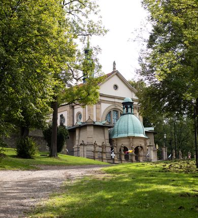 Zdjęcie nr 17: Kościół należy do zespołu dróżek Matki Boskiej, położnych w obrębie klasztoru w Kalwarii Zebrzydowskiej. Świątynia jest położna w centrum wsi, na wschód od głównej drogi, na placu otoczonym kamiennym murem z żeliwnym płotem, który od frontu ozdobiony jest figurami 12 apostołów, umieszczonymi na wysokich cokołach. Główne wejście na plac prowadzi przez bramę od zachodu, flankowaną dwoma filarami, na których ustawione są klasycystyczne wazony. Drugie wejście usytuowane jest od wschodu przez murowaną bramę, nakrytą daszkiem dwuspadowym, na którym ustawiona jest figura grającego na trąbie anioła. Trzecie wejście znajduje się od północy.

Kościół jest budowlą orientowaną, założoną na planie prostokąta, jednonawową, zamkniętą półkolistą absydą. Od zachodu przylega kruchta na planie prostokąta, zamkniętego z jednej strony półkoliście. Wewnątrz kościół dwupiętrowy. Na parterze część wschodnia świątyni podniesiona jest o siedem stopni, oddzielona od nawy trzema półkolistymi arkadami, wspartymi na czterech filarach i ustawionymi przed nimi półkolumnami, które wspierają pełne belkowanie, wyłamane na osiach. W przyłuczach płskorzeźbione główki aniołków. Na fryzie złocony napis: „EIA ADVOCATA NOSTRA OCVLOS MISERICORDES AD NOS CONVERTE ET IESVM BENEDICTV FRCTVM NOBIS OSTENDE”. Za środkową arkadą usytuowana jest niewielka, najstarsza część świątyni – Grób Matki Boskiej, na rzucie prostokąta, sklepiony krzyżowo. W ścianach bocznych nawy są półkoliście zamknięte wnęki na ołtarze. Od zachodu chór muzyczny wsparty jest na trzech arkadach filarowych. Wnętrze obiega drewniany ganek wsparty na metalowych wspornikach, a w części wschodniej i zachodniej na trzech murowanych arkadach. Podniebienie ganku zdobione jest geometrycznym stiukiem. W części nawowej świątyni znajduje się sklepienie kolebkowe z lunetami, w apsydzie hemisferyczne. Wnętrze doświetlone jest sześcioma małymi, okrągłymi oknami znajdującymi się na piętrze od strony północnej i południowej.

Fasada w dolnej części zaakcentowana jest przez monumentalną kruchtę w formie aediculi, założoną na rzucie prostokąta zamkniętego półkoliście. Ściany artykułowane są pięcioma półkolistymi arkadami, wspartymi na filarkach. Od zewnątrz osie ujęte są przez kanelowane kolumny, ustawione na wysokich cokołach, na tle pilastrów. Podpory wznoszą pełne belkowanie i wysunięty gzyms zdobiony kimationem i dekoracją kostkową. Nakrycie w formie kopuły półkolistej z latarnią. Belkowanie ozdobione jest dekoracją roślinną i główkami aniołków. Po bokach kruchty wysunięte są fragmenty muru oporowego do połowy wysokości fasady. Powyżej fasada flankowana przez dwie pary pilastrów z jońskimi kapitelami wspierającymi niepełne belkowanie. Pośrodku duże, półkoliste okno z metalową, dekoracyjną kratą. Szczyt trójkątny z oculusem pośrodku. Na szczycie ustawiona jest figura Najświętszej Marii Panny. Świątynia dookoła obwiedziona szerokim murem oporowym, nakrytym daszkiem, o ukośnych ścianach, flankowanych szkarpami, po sześć na każdej ścianie bocznej i dwoma na ścianie zachodniej oraz wschodniej. Powyżej elewacje ujęte pilastrami z kapitelami jońskimi wznoszącymi niepełne belkowanie. Na wschodniej ścianie półkolista apsyda, nakryta półkopułą. Kościół nakryty dachem siodłowym ze zdwojoną, wysoką latarnią pośrodku. Na narożach dachu ustawione wysokie obeliski na cokołach. 

 