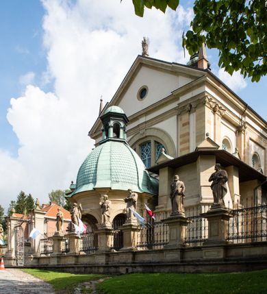 Zdjęcie nr 4: Kościół należy do zespołu dróżek Matki Boskiej, położnych w obrębie klasztoru w Kalwarii Zebrzydowskiej. Świątynia jest położna w centrum wsi, na wschód od głównej drogi, na placu otoczonym kamiennym murem z żeliwnym płotem, który od frontu ozdobiony jest figurami 12 apostołów, umieszczonymi na wysokich cokołach. Główne wejście na plac prowadzi przez bramę od zachodu, flankowaną dwoma filarami, na których ustawione są klasycystyczne wazony. Drugie wejście usytuowane jest od wschodu przez murowaną bramę, nakrytą daszkiem dwuspadowym, na którym ustawiona jest figura grającego na trąbie anioła. Trzecie wejście znajduje się od północy.

Kościół jest budowlą orientowaną, założoną na planie prostokąta, jednonawową, zamkniętą półkolistą absydą. Od zachodu przylega kruchta na planie prostokąta, zamkniętego z jednej strony półkoliście. Wewnątrz kościół dwupiętrowy. Na parterze część wschodnia świątyni podniesiona jest o siedem stopni, oddzielona od nawy trzema półkolistymi arkadami, wspartymi na czterech filarach i ustawionymi przed nimi półkolumnami, które wspierają pełne belkowanie, wyłamane na osiach. W przyłuczach płskorzeźbione główki aniołków. Na fryzie złocony napis: „EIA ADVOCATA NOSTRA OCVLOS MISERICORDES AD NOS CONVERTE ET IESVM BENEDICTV FRCTVM NOBIS OSTENDE”. Za środkową arkadą usytuowana jest niewielka, najstarsza część świątyni – Grób Matki Boskiej, na rzucie prostokąta, sklepiony krzyżowo. W ścianach bocznych nawy są półkoliście zamknięte wnęki na ołtarze. Od zachodu chór muzyczny wsparty jest na trzech arkadach filarowych. Wnętrze obiega drewniany ganek wsparty na metalowych wspornikach, a w części wschodniej i zachodniej na trzech murowanych arkadach. Podniebienie ganku zdobione jest geometrycznym stiukiem. W części nawowej świątyni znajduje się sklepienie kolebkowe z lunetami, w apsydzie hemisferyczne. Wnętrze doświetlone jest sześcioma małymi, okrągłymi oknami znajdującymi się na piętrze od strony północnej i południowej.

Fasada w dolnej części zaakcentowana jest przez monumentalną kruchtę w formie aediculi, założoną na rzucie prostokąta zamkniętego półkoliście. Ściany artykułowane są pięcioma półkolistymi arkadami, wspartymi na filarkach. Od zewnątrz osie ujęte są przez kanelowane kolumny, ustawione na wysokich cokołach, na tle pilastrów. Podpory wznoszą pełne belkowanie i wysunięty gzyms zdobiony kimationem i dekoracją kostkową. Nakrycie w formie kopuły półkolistej z latarnią. Belkowanie ozdobione jest dekoracją roślinną i główkami aniołków. Po bokach kruchty wysunięte są fragmenty muru oporowego do połowy wysokości fasady. Powyżej fasada flankowana przez dwie pary pilastrów z jońskimi kapitelami wspierającymi niepełne belkowanie. Pośrodku duże, półkoliste okno z metalową, dekoracyjną kratą. Szczyt trójkątny z oculusem pośrodku. Na szczycie ustawiona jest figura Najświętszej Marii Panny. Świątynia dookoła obwiedziona szerokim murem oporowym, nakrytym daszkiem, o ukośnych ścianach, flankowanych szkarpami, po sześć na każdej ścianie bocznej i dwoma na ścianie zachodniej oraz wschodniej. Powyżej elewacje ujęte pilastrami z kapitelami jońskimi wznoszącymi niepełne belkowanie. Na wschodniej ścianie półkolista apsyda, nakryta półkopułą. Kościół nakryty dachem siodłowym ze zdwojoną, wysoką latarnią pośrodku. Na narożach dachu ustawione wysokie obeliski na cokołach. 

 