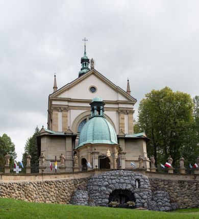 Zdjęcie nr 16: Kościół należy do zespołu dróżek Matki Boskiej, położnych w obrębie klasztoru w Kalwarii Zebrzydowskiej. Świątynia jest położna w centrum wsi, na wschód od głównej drogi, na placu otoczonym kamiennym murem z żeliwnym płotem, który od frontu ozdobiony jest figurami 12 apostołów, umieszczonymi na wysokich cokołach. Główne wejście na plac prowadzi przez bramę od zachodu, flankowaną dwoma filarami, na których ustawione są klasycystyczne wazony. Drugie wejście usytuowane jest od wschodu przez murowaną bramę, nakrytą daszkiem dwuspadowym, na którym ustawiona jest figura grającego na trąbie anioła. Trzecie wejście znajduje się od północy.

Kościół jest budowlą orientowaną, założoną na planie prostokąta, jednonawową, zamkniętą półkolistą absydą. Od zachodu przylega kruchta na planie prostokąta, zamkniętego z jednej strony półkoliście. Wewnątrz kościół dwupiętrowy. Na parterze część wschodnia świątyni podniesiona jest o siedem stopni, oddzielona od nawy trzema półkolistymi arkadami, wspartymi na czterech filarach i ustawionymi przed nimi półkolumnami, które wspierają pełne belkowanie, wyłamane na osiach. W przyłuczach płskorzeźbione główki aniołków. Na fryzie złocony napis: „EIA ADVOCATA NOSTRA OCVLOS MISERICORDES AD NOS CONVERTE ET IESVM BENEDICTV FRCTVM NOBIS OSTENDE”. Za środkową arkadą usytuowana jest niewielka, najstarsza część świątyni – Grób Matki Boskiej, na rzucie prostokąta, sklepiony krzyżowo. W ścianach bocznych nawy są półkoliście zamknięte wnęki na ołtarze. Od zachodu chór muzyczny wsparty jest na trzech arkadach filarowych. Wnętrze obiega drewniany ganek wsparty na metalowych wspornikach, a w części wschodniej i zachodniej na trzech murowanych arkadach. Podniebienie ganku zdobione jest geometrycznym stiukiem. W części nawowej świątyni znajduje się sklepienie kolebkowe z lunetami, w apsydzie hemisferyczne. Wnętrze doświetlone jest sześcioma małymi, okrągłymi oknami znajdującymi się na piętrze od strony północnej i południowej.

Fasada w dolnej części zaakcentowana jest przez monumentalną kruchtę w formie aediculi, założoną na rzucie prostokąta zamkniętego półkoliście. Ściany artykułowane są pięcioma półkolistymi arkadami, wspartymi na filarkach. Od zewnątrz osie ujęte są przez kanelowane kolumny, ustawione na wysokich cokołach, na tle pilastrów. Podpory wznoszą pełne belkowanie i wysunięty gzyms zdobiony kimationem i dekoracją kostkową. Nakrycie w formie kopuły półkolistej z latarnią. Belkowanie ozdobione jest dekoracją roślinną i główkami aniołków. Po bokach kruchty wysunięte są fragmenty muru oporowego do połowy wysokości fasady. Powyżej fasada flankowana przez dwie pary pilastrów z jońskimi kapitelami wspierającymi niepełne belkowanie. Pośrodku duże, półkoliste okno z metalową, dekoracyjną kratą. Szczyt trójkątny z oculusem pośrodku. Na szczycie ustawiona jest figura Najświętszej Marii Panny. Świątynia dookoła obwiedziona szerokim murem oporowym, nakrytym daszkiem, o ukośnych ścianach, flankowanych szkarpami, po sześć na każdej ścianie bocznej i dwoma na ścianie zachodniej oraz wschodniej. Powyżej elewacje ujęte pilastrami z kapitelami jońskimi wznoszącymi niepełne belkowanie. Na wschodniej ścianie półkolista apsyda, nakryta półkopułą. Kościół nakryty dachem siodłowym ze zdwojoną, wysoką latarnią pośrodku. Na narożach dachu ustawione wysokie obeliski na cokołach. 

 