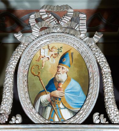 Zdjęcie nr 9: Ołtarz ustawiony na piętrze kościoła. Nastawa jednokondygnacyjna, trójosiowa, ażurowa z dostawionymi po bokach małymi ołtarzykami. Tabernakulum w formie prostopadłościennej skrzynki o wykroju wklęsło-wypukłym, ustawione na cokole i zamknięte profilowanym, wyłamanym gzymsem. Ścianki boczne ustawione pod skosem, wklęsłe, zdobione ukośną, ażurową dekoracją ornamentalną składającą się z kratki, wolut i liści akantu. Ścianka frontowa, wypukła flankowana parą ukośnie ustawionych pilastrów, których trzony zdobione są spływami wolutowymi oraz kwiatowymi. Pośrodku wnęka z metalowym, ruchomym i obrotowym (?) wsadem, który na jednej ze ścianek zdobiony jest płyciną w kształcie stojącego prostokąta zamkniętego łukiem wklęsło-wypukłym z uskokami z przedstawieniem Baranka Bożego na księdze zamkniętej siedmioma pieczęciami, liter alfa i omega oraz powyżej gołębicy Ducha Świętego. Płycina ujęta kimationem, w przyłuczach winne grona. 

Nastawa wsparta na wysokim cokole. Oś główna flankowana parą krótkich pilastrów o kanelowanych trzonach i kompozytowych kapitelach. Przed nimi ustawiona ukośnie para kolumn o dłuższych trzonach, wspartych na pojedynczych, oddzielnych cokołach. Podpory wspólnie wspierają przerwane belkowanie. Kolumny mają bazy, kanelowane trzony i kompozytowe kapitele. W polu głównym znajduje się głęboka wnęka ujęta szeroką, dekoracyjną ramą w kształcie stojącego prostokąta z uszakami, zamknięta łukiem dwuramiennym. Wewnątrz ustawiona jest figura Matki Boskiej Niepokalanie Poczętej na czerwonym materiałowym tle. Dokoła wnęki bogata dekoracja utworzona z gałązek róż i obłoków. Nad wnęką okrągły medalion, ozdobiony u góry karbowaną wstęgą z napisem: „«Astitit / Regina a dex»/Tris tuis in ve/Stitu de[a]vrato / Psal 44 ver 11”. Pomiędzy osią główną, a flankującymi ją pilastrami prześwity. 

Zwieńczenie w formie monumentalnej glorii z wpisanym w polu otokiem z obłoków. Wewnątrz rzeźbiona Trójca Święta oraz cztery uskrzydlone główki anielskie. Na belkowaniu usadzone dwie pary muzykujących aniołów oraz jedna para wysokich wazonów z bujnymi kwiatami. 

W osiach bocznych, w dolnej części cokołowej bramki ujęte szerokimi, kanelowanymi pilastrami podtrzymującymi belkowanie. Przejścia w kształcie stojących prostokątów, zamkniętych półkoliście, flankowane filarkami. W górnej części osi bocznych na niskim podeście ustawione są wysokie, wąskie kanelowane filary ze stylizowanymi jońskimi kapitelami, na których na postumentach w kształcie stylizowanej egipskiej głowicy ustawione są figury: króla Saula z małym Dawidem w lewej osi oraz króla Dawida w prawej osi bocznej. Filary ujęte są po bokach ażurową snycerską dekoracją złożoną z esownic zdobionych cekinami oraz dekoracji roślinnej utworzonej z liści stylizowanego akantu oraz kwiatów. 

Po bokach ołtarza dostawiona jest para małych ołtarzyków, jednokondygnacyjnych i jednoosiowych. Mensy sarkofagowe o kształcie niesymetrycznym: prawy bok przylega do ściany; w polu dwie fornirowane płyciny. Nastawy ołtarzowe, symetryczne, wsparte na łamanym cokole, ujęte parą pilastrów oraz parą kolumn z kanelowanymi i złoconymi trzonami oraz o kompozytowych kapitelach. Podpory wspierają wyłamane belkowania i silnie profilowane gzymsy. Ołtarze zwieńczone są promienistymi gloriami, w których w niebieskim polu, otoczonym obłokami znajduje się: w ołtarzyku po lewej stornie kwiat róży, a w ołtarzyku po prawej stronie gwiazda. W polach centralnych w ramach w kształcie stojących prostokątów z uszakami o wklęsłych górnych narożach umieszczone są obrazy. W ołtarzyku po lewej stronie obraz przedstawiający „Pokłon pasterzy”, a w ołtarzyku po prawej stronie obraz „Pokłon Trzech Króli”. 

Struktura polichromowana w kolorze jasnoniebieskim; ornamentyka, detale i profilowania złocone oraz srebrzone. 


