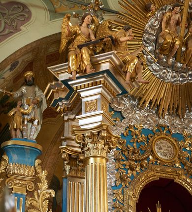 Zdjęcie nr 6: Ołtarz ustawiony na piętrze kościoła. Nastawa jednokondygnacyjna, trójosiowa, ażurowa z dostawionymi po bokach małymi ołtarzykami. Tabernakulum w formie prostopadłościennej skrzynki o wykroju wklęsło-wypukłym, ustawione na cokole i zamknięte profilowanym, wyłamanym gzymsem. Ścianki boczne ustawione pod skosem, wklęsłe, zdobione ukośną, ażurową dekoracją ornamentalną składającą się z kratki, wolut i liści akantu. Ścianka frontowa, wypukła flankowana parą ukośnie ustawionych pilastrów, których trzony zdobione są spływami wolutowymi oraz kwiatowymi. Pośrodku wnęka z metalowym, ruchomym i obrotowym (?) wsadem, który na jednej ze ścianek zdobiony jest płyciną w kształcie stojącego prostokąta zamkniętego łukiem wklęsło-wypukłym z uskokami z przedstawieniem Baranka Bożego na księdze zamkniętej siedmioma pieczęciami, liter alfa i omega oraz powyżej gołębicy Ducha Świętego. Płycina ujęta kimationem, w przyłuczach winne grona. 

Nastawa wsparta na wysokim cokole. Oś główna flankowana parą krótkich pilastrów o kanelowanych trzonach i kompozytowych kapitelach. Przed nimi ustawiona ukośnie para kolumn o dłuższych trzonach, wspartych na pojedynczych, oddzielnych cokołach. Podpory wspólnie wspierają przerwane belkowanie. Kolumny mają bazy, kanelowane trzony i kompozytowe kapitele. W polu głównym znajduje się głęboka wnęka ujęta szeroką, dekoracyjną ramą w kształcie stojącego prostokąta z uszakami, zamknięta łukiem dwuramiennym. Wewnątrz ustawiona jest figura Matki Boskiej Niepokalanie Poczętej na czerwonym materiałowym tle. Dokoła wnęki bogata dekoracja utworzona z gałązek róż i obłoków. Nad wnęką okrągły medalion, ozdobiony u góry karbowaną wstęgą z napisem: „«Astitit / Regina a dex»/Tris tuis in ve/Stitu de[a]vrato / Psal 44 ver 11”. Pomiędzy osią główną, a flankującymi ją pilastrami prześwity. 

Zwieńczenie w formie monumentalnej glorii z wpisanym w polu otokiem z obłoków. Wewnątrz rzeźbiona Trójca Święta oraz cztery uskrzydlone główki anielskie. Na belkowaniu usadzone dwie pary muzykujących aniołów oraz jedna para wysokich wazonów z bujnymi kwiatami. 

W osiach bocznych, w dolnej części cokołowej bramki ujęte szerokimi, kanelowanymi pilastrami podtrzymującymi belkowanie. Przejścia w kształcie stojących prostokątów, zamkniętych półkoliście, flankowane filarkami. W górnej części osi bocznych na niskim podeście ustawione są wysokie, wąskie kanelowane filary ze stylizowanymi jońskimi kapitelami, na których na postumentach w kształcie stylizowanej egipskiej głowicy ustawione są figury: króla Saula z małym Dawidem w lewej osi oraz króla Dawida w prawej osi bocznej. Filary ujęte są po bokach ażurową snycerską dekoracją złożoną z esownic zdobionych cekinami oraz dekoracji roślinnej utworzonej z liści stylizowanego akantu oraz kwiatów. 

Po bokach ołtarza dostawiona jest para małych ołtarzyków, jednokondygnacyjnych i jednoosiowych. Mensy sarkofagowe o kształcie niesymetrycznym: prawy bok przylega do ściany; w polu dwie fornirowane płyciny. Nastawy ołtarzowe, symetryczne, wsparte na łamanym cokole, ujęte parą pilastrów oraz parą kolumn z kanelowanymi i złoconymi trzonami oraz o kompozytowych kapitelach. Podpory wspierają wyłamane belkowania i silnie profilowane gzymsy. Ołtarze zwieńczone są promienistymi gloriami, w których w niebieskim polu, otoczonym obłokami znajduje się: w ołtarzyku po lewej stornie kwiat róży, a w ołtarzyku po prawej stronie gwiazda. W polach centralnych w ramach w kształcie stojących prostokątów z uszakami o wklęsłych górnych narożach umieszczone są obrazy. W ołtarzyku po lewej stronie obraz przedstawiający „Pokłon pasterzy”, a w ołtarzyku po prawej stronie obraz „Pokłon Trzech Króli”. 

Struktura polichromowana w kolorze jasnoniebieskim; ornamentyka, detale i profilowania złocone oraz srebrzone. 


