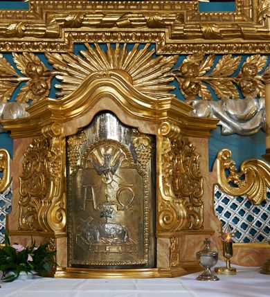 Zdjęcie nr 7: Ołtarz ustawiony na piętrze kościoła. Nastawa jednokondygnacyjna, trójosiowa, ażurowa z dostawionymi po bokach małymi ołtarzykami. Tabernakulum w formie prostopadłościennej skrzynki o wykroju wklęsło-wypukłym, ustawione na cokole i zamknięte profilowanym, wyłamanym gzymsem. Ścianki boczne ustawione pod skosem, wklęsłe, zdobione ukośną, ażurową dekoracją ornamentalną składającą się z kratki, wolut i liści akantu. Ścianka frontowa, wypukła flankowana parą ukośnie ustawionych pilastrów, których trzony zdobione są spływami wolutowymi oraz kwiatowymi. Pośrodku wnęka z metalowym, ruchomym i obrotowym (?) wsadem, który na jednej ze ścianek zdobiony jest płyciną w kształcie stojącego prostokąta zamkniętego łukiem wklęsło-wypukłym z uskokami z przedstawieniem Baranka Bożego na księdze zamkniętej siedmioma pieczęciami, liter alfa i omega oraz powyżej gołębicy Ducha Świętego. Płycina ujęta kimationem, w przyłuczach winne grona. 

Nastawa wsparta na wysokim cokole. Oś główna flankowana parą krótkich pilastrów o kanelowanych trzonach i kompozytowych kapitelach. Przed nimi ustawiona ukośnie para kolumn o dłuższych trzonach, wspartych na pojedynczych, oddzielnych cokołach. Podpory wspólnie wspierają przerwane belkowanie. Kolumny mają bazy, kanelowane trzony i kompozytowe kapitele. W polu głównym znajduje się głęboka wnęka ujęta szeroką, dekoracyjną ramą w kształcie stojącego prostokąta z uszakami, zamknięta łukiem dwuramiennym. Wewnątrz ustawiona jest figura Matki Boskiej Niepokalanie Poczętej na czerwonym materiałowym tle. Dokoła wnęki bogata dekoracja utworzona z gałązek róż i obłoków. Nad wnęką okrągły medalion, ozdobiony u góry karbowaną wstęgą z napisem: „«Astitit / Regina a dex»/Tris tuis in ve/Stitu de[a]vrato / Psal 44 ver 11”. Pomiędzy osią główną, a flankującymi ją pilastrami prześwity. 

Zwieńczenie w formie monumentalnej glorii z wpisanym w polu otokiem z obłoków. Wewnątrz rzeźbiona Trójca Święta oraz cztery uskrzydlone główki anielskie. Na belkowaniu usadzone dwie pary muzykujących aniołów oraz jedna para wysokich wazonów z bujnymi kwiatami. 

W osiach bocznych, w dolnej części cokołowej bramki ujęte szerokimi, kanelowanymi pilastrami podtrzymującymi belkowanie. Przejścia w kształcie stojących prostokątów, zamkniętych półkoliście, flankowane filarkami. W górnej części osi bocznych na niskim podeście ustawione są wysokie, wąskie kanelowane filary ze stylizowanymi jońskimi kapitelami, na których na postumentach w kształcie stylizowanej egipskiej głowicy ustawione są figury: króla Saula z małym Dawidem w lewej osi oraz króla Dawida w prawej osi bocznej. Filary ujęte są po bokach ażurową snycerską dekoracją złożoną z esownic zdobionych cekinami oraz dekoracji roślinnej utworzonej z liści stylizowanego akantu oraz kwiatów. 

Po bokach ołtarza dostawiona jest para małych ołtarzyków, jednokondygnacyjnych i jednoosiowych. Mensy sarkofagowe o kształcie niesymetrycznym: prawy bok przylega do ściany; w polu dwie fornirowane płyciny. Nastawy ołtarzowe, symetryczne, wsparte na łamanym cokole, ujęte parą pilastrów oraz parą kolumn z kanelowanymi i złoconymi trzonami oraz o kompozytowych kapitelach. Podpory wspierają wyłamane belkowania i silnie profilowane gzymsy. Ołtarze zwieńczone są promienistymi gloriami, w których w niebieskim polu, otoczonym obłokami znajduje się: w ołtarzyku po lewej stornie kwiat róży, a w ołtarzyku po prawej stronie gwiazda. W polach centralnych w ramach w kształcie stojących prostokątów z uszakami o wklęsłych górnych narożach umieszczone są obrazy. W ołtarzyku po lewej stronie obraz przedstawiający „Pokłon pasterzy”, a w ołtarzyku po prawej stronie obraz „Pokłon Trzech Króli”. 

Struktura polichromowana w kolorze jasnoniebieskim; ornamentyka, detale i profilowania złocone oraz srebrzone. 


