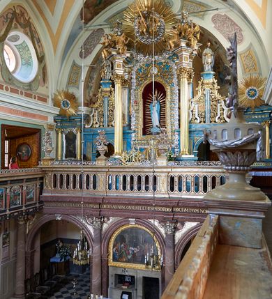 Zdjęcie nr 3: Ołtarz ustawiony na piętrze kościoła. Nastawa jednokondygnacyjna, trójosiowa, ażurowa z dostawionymi po bokach małymi ołtarzykami. Tabernakulum w formie prostopadłościennej skrzynki o wykroju wklęsło-wypukłym, ustawione na cokole i zamknięte profilowanym, wyłamanym gzymsem. Ścianki boczne ustawione pod skosem, wklęsłe, zdobione ukośną, ażurową dekoracją ornamentalną składającą się z kratki, wolut i liści akantu. Ścianka frontowa, wypukła flankowana parą ukośnie ustawionych pilastrów, których trzony zdobione są spływami wolutowymi oraz kwiatowymi. Pośrodku wnęka z metalowym, ruchomym i obrotowym (?) wsadem, który na jednej ze ścianek zdobiony jest płyciną w kształcie stojącego prostokąta zamkniętego łukiem wklęsło-wypukłym z uskokami z przedstawieniem Baranka Bożego na księdze zamkniętej siedmioma pieczęciami, liter alfa i omega oraz powyżej gołębicy Ducha Świętego. Płycina ujęta kimationem, w przyłuczach winne grona. 

Nastawa wsparta na wysokim cokole. Oś główna flankowana parą krótkich pilastrów o kanelowanych trzonach i kompozytowych kapitelach. Przed nimi ustawiona ukośnie para kolumn o dłuższych trzonach, wspartych na pojedynczych, oddzielnych cokołach. Podpory wspólnie wspierają przerwane belkowanie. Kolumny mają bazy, kanelowane trzony i kompozytowe kapitele. W polu głównym znajduje się głęboka wnęka ujęta szeroką, dekoracyjną ramą w kształcie stojącego prostokąta z uszakami, zamknięta łukiem dwuramiennym. Wewnątrz ustawiona jest figura Matki Boskiej Niepokalanie Poczętej na czerwonym materiałowym tle. Dokoła wnęki bogata dekoracja utworzona z gałązek róż i obłoków. Nad wnęką okrągły medalion, ozdobiony u góry karbowaną wstęgą z napisem: „«Astitit / Regina a dex»/Tris tuis in ve/Stitu de[a]vrato / Psal 44 ver 11”. Pomiędzy osią główną, a flankującymi ją pilastrami prześwity. 

Zwieńczenie w formie monumentalnej glorii z wpisanym w polu otokiem z obłoków. Wewnątrz rzeźbiona Trójca Święta oraz cztery uskrzydlone główki anielskie. Na belkowaniu usadzone dwie pary muzykujących aniołów oraz jedna para wysokich wazonów z bujnymi kwiatami. 

W osiach bocznych, w dolnej części cokołowej bramki ujęte szerokimi, kanelowanymi pilastrami podtrzymującymi belkowanie. Przejścia w kształcie stojących prostokątów, zamkniętych półkoliście, flankowane filarkami. W górnej części osi bocznych na niskim podeście ustawione są wysokie, wąskie kanelowane filary ze stylizowanymi jońskimi kapitelami, na których na postumentach w kształcie stylizowanej egipskiej głowicy ustawione są figury: króla Saula z małym Dawidem w lewej osi oraz króla Dawida w prawej osi bocznej. Filary ujęte są po bokach ażurową snycerską dekoracją złożoną z esownic zdobionych cekinami oraz dekoracji roślinnej utworzonej z liści stylizowanego akantu oraz kwiatów. 

Po bokach ołtarza dostawiona jest para małych ołtarzyków, jednokondygnacyjnych i jednoosiowych. Mensy sarkofagowe o kształcie niesymetrycznym: prawy bok przylega do ściany; w polu dwie fornirowane płyciny. Nastawy ołtarzowe, symetryczne, wsparte na łamanym cokole, ujęte parą pilastrów oraz parą kolumn z kanelowanymi i złoconymi trzonami oraz o kompozytowych kapitelach. Podpory wspierają wyłamane belkowania i silnie profilowane gzymsy. Ołtarze zwieńczone są promienistymi gloriami, w których w niebieskim polu, otoczonym obłokami znajduje się: w ołtarzyku po lewej stornie kwiat róży, a w ołtarzyku po prawej stronie gwiazda. W polach centralnych w ramach w kształcie stojących prostokątów z uszakami o wklęsłych górnych narożach umieszczone są obrazy. W ołtarzyku po lewej stronie obraz przedstawiający „Pokłon pasterzy”, a w ołtarzyku po prawej stronie obraz „Pokłon Trzech Króli”. 

Struktura polichromowana w kolorze jasnoniebieskim; ornamentyka, detale i profilowania złocone oraz srebrzone. 


