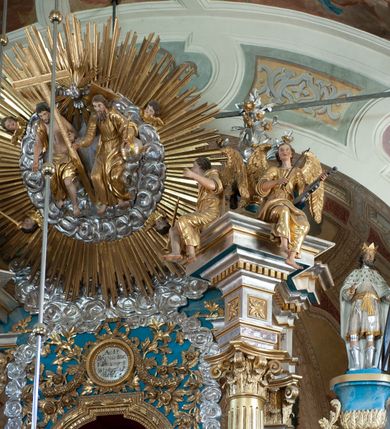 Zdjęcie nr 5: Ołtarz ustawiony na piętrze kościoła. Nastawa jednokondygnacyjna, trójosiowa, ażurowa z dostawionymi po bokach małymi ołtarzykami. Tabernakulum w formie prostopadłościennej skrzynki o wykroju wklęsło-wypukłym, ustawione na cokole i zamknięte profilowanym, wyłamanym gzymsem. Ścianki boczne ustawione pod skosem, wklęsłe, zdobione ukośną, ażurową dekoracją ornamentalną składającą się z kratki, wolut i liści akantu. Ścianka frontowa, wypukła flankowana parą ukośnie ustawionych pilastrów, których trzony zdobione są spływami wolutowymi oraz kwiatowymi. Pośrodku wnęka z metalowym, ruchomym i obrotowym (?) wsadem, który na jednej ze ścianek zdobiony jest płyciną w kształcie stojącego prostokąta zamkniętego łukiem wklęsło-wypukłym z uskokami z przedstawieniem Baranka Bożego na księdze zamkniętej siedmioma pieczęciami, liter alfa i omega oraz powyżej gołębicy Ducha Świętego. Płycina ujęta kimationem, w przyłuczach winne grona. 

Nastawa wsparta na wysokim cokole. Oś główna flankowana parą krótkich pilastrów o kanelowanych trzonach i kompozytowych kapitelach. Przed nimi ustawiona ukośnie para kolumn o dłuższych trzonach, wspartych na pojedynczych, oddzielnych cokołach. Podpory wspólnie wspierają przerwane belkowanie. Kolumny mają bazy, kanelowane trzony i kompozytowe kapitele. W polu głównym znajduje się głęboka wnęka ujęta szeroką, dekoracyjną ramą w kształcie stojącego prostokąta z uszakami, zamknięta łukiem dwuramiennym. Wewnątrz ustawiona jest figura Matki Boskiej Niepokalanie Poczętej na czerwonym materiałowym tle. Dokoła wnęki bogata dekoracja utworzona z gałązek róż i obłoków. Nad wnęką okrągły medalion, ozdobiony u góry karbowaną wstęgą z napisem: „«Astitit / Regina a dex»/Tris tuis in ve/Stitu de[a]vrato / Psal 44 ver 11”. Pomiędzy osią główną, a flankującymi ją pilastrami prześwity. 

Zwieńczenie w formie monumentalnej glorii z wpisanym w polu otokiem z obłoków. Wewnątrz rzeźbiona Trójca Święta oraz cztery uskrzydlone główki anielskie. Na belkowaniu usadzone dwie pary muzykujących aniołów oraz jedna para wysokich wazonów z bujnymi kwiatami. 

W osiach bocznych, w dolnej części cokołowej bramki ujęte szerokimi, kanelowanymi pilastrami podtrzymującymi belkowanie. Przejścia w kształcie stojących prostokątów, zamkniętych półkoliście, flankowane filarkami. W górnej części osi bocznych na niskim podeście ustawione są wysokie, wąskie kanelowane filary ze stylizowanymi jońskimi kapitelami, na których na postumentach w kształcie stylizowanej egipskiej głowicy ustawione są figury: króla Saula z małym Dawidem w lewej osi oraz króla Dawida w prawej osi bocznej. Filary ujęte są po bokach ażurową snycerską dekoracją złożoną z esownic zdobionych cekinami oraz dekoracji roślinnej utworzonej z liści stylizowanego akantu oraz kwiatów. 

Po bokach ołtarza dostawiona jest para małych ołtarzyków, jednokondygnacyjnych i jednoosiowych. Mensy sarkofagowe o kształcie niesymetrycznym: prawy bok przylega do ściany; w polu dwie fornirowane płyciny. Nastawy ołtarzowe, symetryczne, wsparte na łamanym cokole, ujęte parą pilastrów oraz parą kolumn z kanelowanymi i złoconymi trzonami oraz o kompozytowych kapitelach. Podpory wspierają wyłamane belkowania i silnie profilowane gzymsy. Ołtarze zwieńczone są promienistymi gloriami, w których w niebieskim polu, otoczonym obłokami znajduje się: w ołtarzyku po lewej stornie kwiat róży, a w ołtarzyku po prawej stronie gwiazda. W polach centralnych w ramach w kształcie stojących prostokątów z uszakami o wklęsłych górnych narożach umieszczone są obrazy. W ołtarzyku po lewej stronie obraz przedstawiający „Pokłon pasterzy”, a w ołtarzyku po prawej stronie obraz „Pokłon Trzech Króli”. 

Struktura polichromowana w kolorze jasnoniebieskim; ornamentyka, detale i profilowania złocone oraz srebrzone. 


