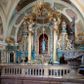 Zdjęcie nr 1: Ołtarz ustawiony na piętrze kościoła. Nastawa jednokondygnacyjna, trójosiowa, ażurowa z dostawionymi po bokach małymi ołtarzykami. Tabernakulum w formie prostopadłościennej skrzynki o wykroju wklęsło-wypukłym, ustawione na cokole i zamknięte profilowanym, wyłamanym gzymsem. Ścianki boczne ustawione pod skosem, wklęsłe, zdobione ukośną, ażurową dekoracją ornamentalną składającą się z kratki, wolut i liści akantu. Ścianka frontowa, wypukła flankowana parą ukośnie ustawionych pilastrów, których trzony zdobione są spływami wolutowymi oraz kwiatowymi. Pośrodku wnęka z metalowym, ruchomym i obrotowym (?) wsadem, który na jednej ze ścianek zdobiony jest płyciną w kształcie stojącego prostokąta zamkniętego łukiem wklęsło-wypukłym z uskokami z przedstawieniem Baranka Bożego na księdze zamkniętej siedmioma pieczęciami, liter alfa i omega oraz powyżej gołębicy Ducha Świętego. Płycina ujęta kimationem, w przyłuczach winne grona. 

Nastawa wsparta na wysokim cokole. Oś główna flankowana parą krótkich pilastrów o kanelowanych trzonach i kompozytowych kapitelach. Przed nimi ustawiona ukośnie para kolumn o dłuższych trzonach, wspartych na pojedynczych, oddzielnych cokołach. Podpory wspólnie wspierają przerwane belkowanie. Kolumny mają bazy, kanelowane trzony i kompozytowe kapitele. W polu głównym znajduje się głęboka wnęka ujęta szeroką, dekoracyjną ramą w kształcie stojącego prostokąta z uszakami, zamknięta łukiem dwuramiennym. Wewnątrz ustawiona jest figura Matki Boskiej Niepokalanie Poczętej na czerwonym materiałowym tle. Dokoła wnęki bogata dekoracja utworzona z gałązek róż i obłoków. Nad wnęką okrągły medalion, ozdobiony u góry karbowaną wstęgą z napisem: „«Astitit / Regina a dex»/Tris tuis in ve/Stitu de[a]vrato / Psal 44 ver 11”. Pomiędzy osią główną, a flankującymi ją pilastrami prześwity. 

Zwieńczenie w formie monumentalnej glorii z wpisanym w polu otokiem z obłoków. Wewnątrz rzeźbiona Trójca Święta oraz cztery uskrzydlone główki anielskie. Na belkowaniu usadzone dwie pary muzykujących aniołów oraz jedna para wysokich wazonów z bujnymi kwiatami. 

W osiach bocznych, w dolnej części cokołowej bramki ujęte szerokimi, kanelowanymi pilastrami podtrzymującymi belkowanie. Przejścia w kształcie stojących prostokątów, zamkniętych półkoliście, flankowane filarkami. W górnej części osi bocznych na niskim podeście ustawione są wysokie, wąskie kanelowane filary ze stylizowanymi jońskimi kapitelami, na których na postumentach w kształcie stylizowanej egipskiej głowicy ustawione są figury: króla Saula z małym Dawidem w lewej osi oraz króla Dawida w prawej osi bocznej. Filary ujęte są po bokach ażurową snycerską dekoracją złożoną z esownic zdobionych cekinami oraz dekoracji roślinnej utworzonej z liści stylizowanego akantu oraz kwiatów. 

Po bokach ołtarza dostawiona jest para małych ołtarzyków, jednokondygnacyjnych i jednoosiowych. Mensy sarkofagowe o kształcie niesymetrycznym: prawy bok przylega do ściany; w polu dwie fornirowane płyciny. Nastawy ołtarzowe, symetryczne, wsparte na łamanym cokole, ujęte parą pilastrów oraz parą kolumn z kanelowanymi i złoconymi trzonami oraz o kompozytowych kapitelach. Podpory wspierają wyłamane belkowania i silnie profilowane gzymsy. Ołtarze zwieńczone są promienistymi gloriami, w których w niebieskim polu, otoczonym obłokami znajduje się: w ołtarzyku po lewej stornie kwiat róży, a w ołtarzyku po prawej stronie gwiazda. W polach centralnych w ramach w kształcie stojących prostokątów z uszakami o wklęsłych górnych narożach umieszczone są obrazy. W ołtarzyku po lewej stronie obraz przedstawiający „Pokłon pasterzy”, a w ołtarzyku po prawej stronie obraz „Pokłon Trzech Króli”. 

Struktura polichromowana w kolorze jasnoniebieskim; ornamentyka, detale i profilowania złocone oraz srebrzone. 


