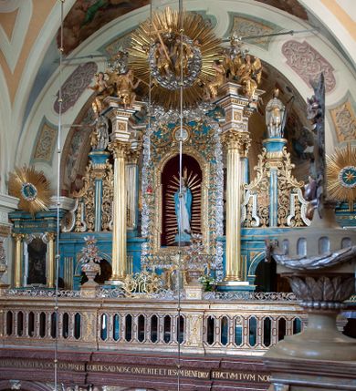 Zdjęcie nr 1: Ołtarz ustawiony na piętrze kościoła. Nastawa jednokondygnacyjna, trójosiowa, ażurowa z dostawionymi po bokach małymi ołtarzykami. Tabernakulum w formie prostopadłościennej skrzynki o wykroju wklęsło-wypukłym, ustawione na cokole i zamknięte profilowanym, wyłamanym gzymsem. Ścianki boczne ustawione pod skosem, wklęsłe, zdobione ukośną, ażurową dekoracją ornamentalną składającą się z kratki, wolut i liści akantu. Ścianka frontowa, wypukła flankowana parą ukośnie ustawionych pilastrów, których trzony zdobione są spływami wolutowymi oraz kwiatowymi. Pośrodku wnęka z metalowym, ruchomym i obrotowym (?) wsadem, który na jednej ze ścianek zdobiony jest płyciną w kształcie stojącego prostokąta zamkniętego łukiem wklęsło-wypukłym z uskokami z przedstawieniem Baranka Bożego na księdze zamkniętej siedmioma pieczęciami, liter alfa i omega oraz powyżej gołębicy Ducha Świętego. Płycina ujęta kimationem, w przyłuczach winne grona. 

Nastawa wsparta na wysokim cokole. Oś główna flankowana parą krótkich pilastrów o kanelowanych trzonach i kompozytowych kapitelach. Przed nimi ustawiona ukośnie para kolumn o dłuższych trzonach, wspartych na pojedynczych, oddzielnych cokołach. Podpory wspólnie wspierają przerwane belkowanie. Kolumny mają bazy, kanelowane trzony i kompozytowe kapitele. W polu głównym znajduje się głęboka wnęka ujęta szeroką, dekoracyjną ramą w kształcie stojącego prostokąta z uszakami, zamknięta łukiem dwuramiennym. Wewnątrz ustawiona jest figura Matki Boskiej Niepokalanie Poczętej na czerwonym materiałowym tle. Dokoła wnęki bogata dekoracja utworzona z gałązek róż i obłoków. Nad wnęką okrągły medalion, ozdobiony u góry karbowaną wstęgą z napisem: „«Astitit / Regina a dex»/Tris tuis in ve/Stitu de[a]vrato / Psal 44 ver 11”. Pomiędzy osią główną, a flankującymi ją pilastrami prześwity. 

Zwieńczenie w formie monumentalnej glorii z wpisanym w polu otokiem z obłoków. Wewnątrz rzeźbiona Trójca Święta oraz cztery uskrzydlone główki anielskie. Na belkowaniu usadzone dwie pary muzykujących aniołów oraz jedna para wysokich wazonów z bujnymi kwiatami. 

W osiach bocznych, w dolnej części cokołowej bramki ujęte szerokimi, kanelowanymi pilastrami podtrzymującymi belkowanie. Przejścia w kształcie stojących prostokątów, zamkniętych półkoliście, flankowane filarkami. W górnej części osi bocznych na niskim podeście ustawione są wysokie, wąskie kanelowane filary ze stylizowanymi jońskimi kapitelami, na których na postumentach w kształcie stylizowanej egipskiej głowicy ustawione są figury: króla Saula z małym Dawidem w lewej osi oraz króla Dawida w prawej osi bocznej. Filary ujęte są po bokach ażurową snycerską dekoracją złożoną z esownic zdobionych cekinami oraz dekoracji roślinnej utworzonej z liści stylizowanego akantu oraz kwiatów. 

Po bokach ołtarza dostawiona jest para małych ołtarzyków, jednokondygnacyjnych i jednoosiowych. Mensy sarkofagowe o kształcie niesymetrycznym: prawy bok przylega do ściany; w polu dwie fornirowane płyciny. Nastawy ołtarzowe, symetryczne, wsparte na łamanym cokole, ujęte parą pilastrów oraz parą kolumn z kanelowanymi i złoconymi trzonami oraz o kompozytowych kapitelach. Podpory wspierają wyłamane belkowania i silnie profilowane gzymsy. Ołtarze zwieńczone są promienistymi gloriami, w których w niebieskim polu, otoczonym obłokami znajduje się: w ołtarzyku po lewej stornie kwiat róży, a w ołtarzyku po prawej stronie gwiazda. W polach centralnych w ramach w kształcie stojących prostokątów z uszakami o wklęsłych górnych narożach umieszczone są obrazy. W ołtarzyku po lewej stronie obraz przedstawiający „Pokłon pasterzy”, a w ołtarzyku po prawej stronie obraz „Pokłon Trzech Króli”. 

Struktura polichromowana w kolorze jasnoniebieskim; ornamentyka, detale i profilowania złocone oraz srebrzone. 


