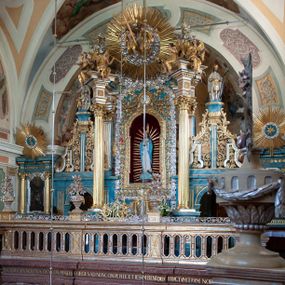 Zdjęcie nr 1: Ołtarz ustawiony na piętrze kościoła. Nastawa jednokondygnacyjna, trójosiowa, ażurowa z dostawionymi po bokach małymi ołtarzykami. Tabernakulum w formie prostopadłościennej skrzynki o wykroju wklęsło-wypukłym, ustawione na cokole i zamknięte profilowanym, wyłamanym gzymsem. Ścianki boczne ustawione pod skosem, wklęsłe, zdobione ukośną, ażurową dekoracją ornamentalną składającą się z kratki, wolut i liści akantu. Ścianka frontowa, wypukła flankowana parą ukośnie ustawionych pilastrów, których trzony zdobione są spływami wolutowymi oraz kwiatowymi. Pośrodku wnęka z metalowym, ruchomym i obrotowym (?) wsadem, który na jednej ze ścianek zdobiony jest płyciną w kształcie stojącego prostokąta zamkniętego łukiem wklęsło-wypukłym z uskokami z przedstawieniem Baranka Bożego na księdze zamkniętej siedmioma pieczęciami, liter alfa i omega oraz powyżej gołębicy Ducha Świętego. Płycina ujęta kimationem, w przyłuczach winne grona. 

Nastawa wsparta na wysokim cokole. Oś główna flankowana parą krótkich pilastrów o kanelowanych trzonach i kompozytowych kapitelach. Przed nimi ustawiona ukośnie para kolumn o dłuższych trzonach, wspartych na pojedynczych, oddzielnych cokołach. Podpory wspólnie wspierają przerwane belkowanie. Kolumny mają bazy, kanelowane trzony i kompozytowe kapitele. W polu głównym znajduje się głęboka wnęka ujęta szeroką, dekoracyjną ramą w kształcie stojącego prostokąta z uszakami, zamknięta łukiem dwuramiennym. Wewnątrz ustawiona jest figura Matki Boskiej Niepokalanie Poczętej na czerwonym materiałowym tle. Dokoła wnęki bogata dekoracja utworzona z gałązek róż i obłoków. Nad wnęką okrągły medalion, ozdobiony u góry karbowaną wstęgą z napisem: „«Astitit / Regina a dex»/Tris tuis in ve/Stitu de[a]vrato / Psal 44 ver 11”. Pomiędzy osią główną, a flankującymi ją pilastrami prześwity. 

Zwieńczenie w formie monumentalnej glorii z wpisanym w polu otokiem z obłoków. Wewnątrz rzeźbiona Trójca Święta oraz cztery uskrzydlone główki anielskie. Na belkowaniu usadzone dwie pary muzykujących aniołów oraz jedna para wysokich wazonów z bujnymi kwiatami. 

W osiach bocznych, w dolnej części cokołowej bramki ujęte szerokimi, kanelowanymi pilastrami podtrzymującymi belkowanie. Przejścia w kształcie stojących prostokątów, zamkniętych półkoliście, flankowane filarkami. W górnej części osi bocznych na niskim podeście ustawione są wysokie, wąskie kanelowane filary ze stylizowanymi jońskimi kapitelami, na których na postumentach w kształcie stylizowanej egipskiej głowicy ustawione są figury: króla Saula z małym Dawidem w lewej osi oraz króla Dawida w prawej osi bocznej. Filary ujęte są po bokach ażurową snycerską dekoracją złożoną z esownic zdobionych cekinami oraz dekoracji roślinnej utworzonej z liści stylizowanego akantu oraz kwiatów. 

Po bokach ołtarza dostawiona jest para małych ołtarzyków, jednokondygnacyjnych i jednoosiowych. Mensy sarkofagowe o kształcie niesymetrycznym: prawy bok przylega do ściany; w polu dwie fornirowane płyciny. Nastawy ołtarzowe, symetryczne, wsparte na łamanym cokole, ujęte parą pilastrów oraz parą kolumn z kanelowanymi i złoconymi trzonami oraz o kompozytowych kapitelach. Podpory wspierają wyłamane belkowania i silnie profilowane gzymsy. Ołtarze zwieńczone są promienistymi gloriami, w których w niebieskim polu, otoczonym obłokami znajduje się: w ołtarzyku po lewej stornie kwiat róży, a w ołtarzyku po prawej stronie gwiazda. W polach centralnych w ramach w kształcie stojących prostokątów z uszakami o wklęsłych górnych narożach umieszczone są obrazy. W ołtarzyku po lewej stronie obraz przedstawiający „Pokłon pasterzy”, a w ołtarzyku po prawej stronie obraz „Pokłon Trzech Króli”. 

Struktura polichromowana w kolorze jasnoniebieskim; ornamentyka, detale i profilowania złocone oraz srebrzone. 


