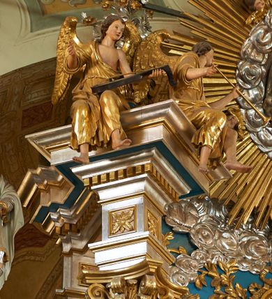 Zdjęcie nr 1: Pełnoplastyczna figura uskrzydlonego anioła ukazanego w pozycji siedzącej, z torsem i głową zwróconymi w lewą stronę. Ręce ugięte, uniesione, w lewej trzyma brązową lutnię. Twarz owalna, pełna, okolona ciemnobrązowymi włosami. Anioł ubrany jest w złoty płaszcz przewieszony przez prawe ramię, odsłaniający lewe ramię. Anioł ma duże złocone skrzydła. Polichromia w odsłoniętych partiach ciała naturalistyczna.
