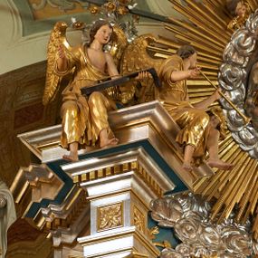 Zdjęcie nr 1: Pełnoplastyczna figura uskrzydlonego anioła ukazanego w pozycji siedzącej, z torsem i głową zwróconymi w lewą stronę. Ręce ugięte, uniesione, w lewej trzyma brązową lutnię. Twarz owalna, pełna, okolona ciemnobrązowymi włosami. Anioł ubrany jest w złoty płaszcz przewieszony przez prawe ramię, odsłaniający lewe ramię. Anioł ma duże złocone skrzydła. Polichromia w odsłoniętych partiach ciała naturalistyczna.
