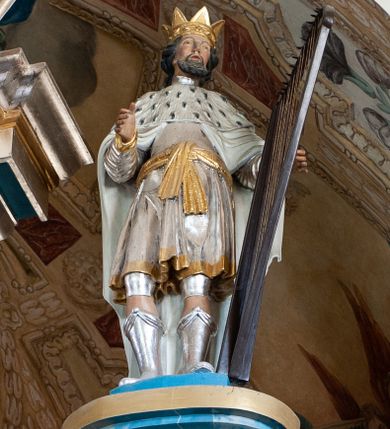 Zdjęcie nr 1: Rzeźba przedstawiająca króla Dawida w postawie stojącej, frontalnie z prawą nogę delikatnie ugiętą w kolanie, z prawą ręką wyciągniętą przed siebie, w lewej trzymającego harfę. Twarz szeroka z długim nosem, delikatnie rozchylonymi ustami, okolona ciemną i krótką brodą oraz średniej długości włosami. Król ubrany jest w srebrną tunikę ze złotą lamówką, przewiązaną złotym pasem oraz biały płaszcz z gronostajową pelerynką, na głowie ma założoną otwartą koronę, a na stopach buty z wysokimi cholewami. Polichromia w odsłoniętych partiach ciała naturalistyczna, detale złocone i srebrzone.


