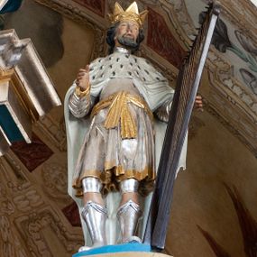 Zdjęcie nr 1: Rzeźba przedstawiająca króla Dawida w postawie stojącej, frontalnie z prawą nogę delikatnie ugiętą w kolanie, z prawą ręką wyciągniętą przed siebie, w lewej trzymającego harfę. Twarz szeroka z długim nosem, delikatnie rozchylonymi ustami, okolona ciemną i krótką brodą oraz średniej długości włosami. Król ubrany jest w srebrną tunikę ze złotą lamówką, przewiązaną złotym pasem oraz biały płaszcz z gronostajową pelerynką, na głowie ma założoną otwartą koronę, a na stopach buty z wysokimi cholewami. Polichromia w odsłoniętych partiach ciała naturalistyczna, detale złocone i srebrzone.


