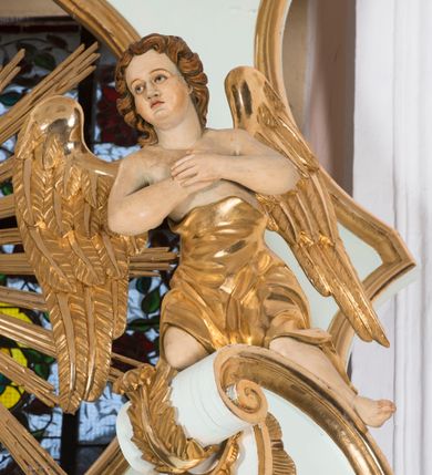 Zdjęcie nr 7: Para pełnoplastycznych, całopostaciowych figur przedstawia anioły przyklękające na przerwanym, wolutowym przyczółku zwieńczenia ołtarza. Ich zwrócone niemal w trzech czwartych, młodzieńcze sylwetki są lekko pochylone ku sobie. Owalne, szerokie twarze aniołów charakteryzują pełne, wyraziste, niemal identyczne rysy z dużymi oczami, długimi, prostymi nosami i lekko zarumienionymi policzkami. Ich falowane, brązowe, częściowo przesłaniające uszy włosy łagodnie opadają na plecy. Sylwetki aniołów, charakteryzujące się stosunkowo naturalistycznie opracowaną muskulaturą, okryte są rozwianymi, złoconymi szatami od pasa do kolan u anioła po prawej stronie i częściowo w partii bioder u anioła po lewej. Na plecach mają duże, złocone skrzydła. Anioł po lewej prawą rękę wspiera na piersiach, lewą wyciąga przed siebie, drugi obydwie składa na klatce piersiowej.      

