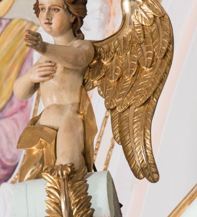 Zdjęcie nr 4: Para pełnoplastycznych, całopostaciowych figur przedstawia anioły przyklękające na przerwanym, wolutowym przyczółku zwieńczenia ołtarza. Ich zwrócone niemal w trzech czwartych, młodzieńcze sylwetki są lekko pochylone ku sobie. Owalne, szerokie twarze aniołów charakteryzują pełne, wyraziste, niemal identyczne rysy z dużymi oczami, długimi, prostymi nosami i lekko zarumienionymi policzkami. Ich falowane, brązowe, częściowo przesłaniające uszy włosy łagodnie opadają na plecy. Sylwetki aniołów, charakteryzujące się stosunkowo naturalistycznie opracowaną muskulaturą, okryte są rozwianymi, złoconymi szatami od pasa do kolan u anioła po prawej stronie i częściowo w partii bioder u anioła po lewej. Na plecach mają duże, złocone skrzydła. Anioł po lewej prawą rękę wspiera na piersiach, lewą wyciąga przed siebie, drugi obydwie składa na klatce piersiowej.      

