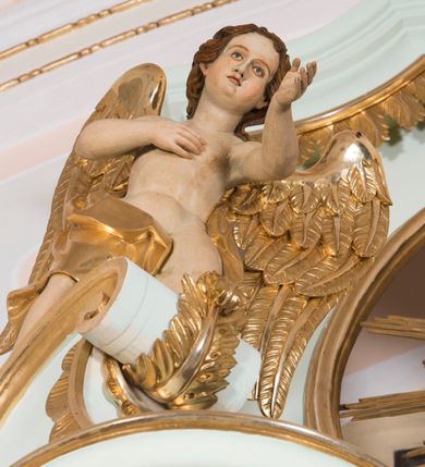 Zdjęcie nr 2: Para pełnoplastycznych, całopostaciowych figur przedstawia anioły przyklękające na przerwanym, wolutowym przyczółku zwieńczenia ołtarza. Ich zwrócone niemal w trzech czwartych, młodzieńcze sylwetki są lekko pochylone ku sobie. Owalne, szerokie twarze aniołów charakteryzują pełne, wyraziste, niemal identyczne rysy z dużymi oczami, długimi, prostymi nosami i lekko zarumienionymi policzkami. Ich falowane, brązowe, częściowo przesłaniające uszy włosy łagodnie opadają na plecy. Sylwetki aniołów, charakteryzujące się stosunkowo naturalistycznie opracowaną muskulaturą, okryte są rozwianymi, złoconymi szatami od pasa do kolan u anioła po prawej stronie i częściowo w partii bioder u anioła po lewej. Na plecach mają duże, złocone skrzydła. Anioł po lewej prawą rękę wspiera na piersiach, lewą wyciąga przed siebie, drugi obydwie składa na klatce piersiowej.      

