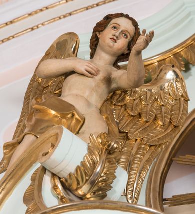 Zdjęcie nr 1: Para pełnoplastycznych, całopostaciowych figur przedstawia anioły przyklękające na przerwanym, wolutowym przyczółku zwieńczenia ołtarza. Ich zwrócone niemal w trzech czwartych, młodzieńcze sylwetki są lekko pochylone ku sobie. Owalne, szerokie twarze aniołów charakteryzują pełne, wyraziste, niemal identyczne rysy z dużymi oczami, długimi, prostymi nosami i lekko zarumienionymi policzkami. Ich falowane, brązowe, częściowo przesłaniające uszy włosy łagodnie opadają na plecy. Sylwetki aniołów, charakteryzujące się stosunkowo naturalistycznie opracowaną muskulaturą, okryte są rozwianymi, złoconymi szatami od pasa do kolan u anioła po prawej stronie i częściowo w partii bioder u anioła po lewej. Na plecach mają duże, złocone skrzydła. Anioł po lewej prawą rękę wspiera na piersiach, lewą wyciąga przed siebie, drugi obydwie składa na klatce piersiowej.      

