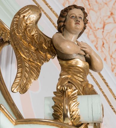 Zdjęcie nr 8: Para pełnoplastycznych, całopostaciowych figur przedstawia anioły przyklękające na przerwanym, wolutowym przyczółku zwieńczenia ołtarza. Ich zwrócone niemal w trzech czwartych, młodzieńcze sylwetki są lekko pochylone ku sobie. Owalne, szerokie twarze aniołów charakteryzują pełne, wyraziste, niemal identyczne rysy z dużymi oczami, długimi, prostymi nosami i lekko zarumienionymi policzkami. Ich falowane, brązowe, częściowo przesłaniające uszy włosy łagodnie opadają na plecy. Sylwetki aniołów, charakteryzujące się stosunkowo naturalistycznie opracowaną muskulaturą, okryte są rozwianymi, złoconymi szatami od pasa do kolan u anioła po prawej stronie i częściowo w partii bioder u anioła po lewej. Na plecach mają duże, złocone skrzydła. Anioł po lewej prawą rękę wspiera na piersiach, lewą wyciąga przed siebie, drugi obydwie składa na klatce piersiowej.      

