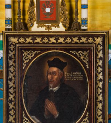 Zdjęcie nr 2: Obraz w kształcie stojącego prostokąta zawiera wizerunek św. Jana Kantego ukazanego w półpostaci, w trzech czwartych zwróconego w prawo. Szczupłą, pociągłą twarz świętego charakteryzują naturalistycznie opracowane rysy z pokrytym drobnymi bruzdami czołem, zwróconymi ku górze, niewielkimi, szeroko osadzonymi oczami, długim, lekko zakrzywionym nosem, wydatnymi kośćmi policzkowymi oraz nieznacznie rozchylonymi ustami widocznymi pomiędzy krótkim, siwym zarostem. Widoczne spod nakrywającego głowę biretu siwe włosy są lekko skręcone i sięgają ramion mężczyzny. Święty Jan Kanty ubrany jest w togę profesorską, ręce ma złożone w geście modlitwy. Głowę świętego okala świetlisty nimb. Postać ukazana jest na ciemnobrązowym, jednolitym tle. Po lewej stronie widoczny jest niewielki krucyfiks, po prawej w głębi grzbiety ksiąg, powyżej, na wysokości głowy świętego znajduje się napis „Effigies S(ancti) IOANṄIS / CANTY post sacrum / è vivis exspiriu(m) deline/ata et depicta, A(nno) D(omini) 1473.”.