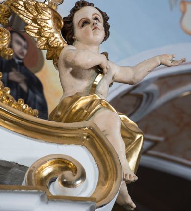 Zdjęcie nr 1: Para pełnoplastycznych, całopostaciowych figur aniołów o sylwetkach dziecięcych przysiadających na spływach wolutowych po obydwu stronach zwieńczenia ołtarza. Zwrócone są one niemal na wprost. Lekko przechylone w bok głowy charakteryzują się zaokrąglonymi twarzami o pełnych, wyrazistych, dziecięcych rysach. Krótkie, kręcone, ciemnobrązowe włosy ujmują twarze zebranymi po bokach i nad czołem puklami. Jedną rękę składają na piersiach, drugą unoszą bądź wyciągają w bok. Nagie sylwetki aniołów osłania w partii bioder złocona tkanina, a przez nagie torsy przewieszone są złocone szarfy. Na plecach mają złocone skrzydełka. 