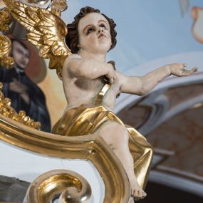 Zdjęcie nr 1: Para pełnoplastycznych, całopostaciowych figur aniołów o sylwetkach dziecięcych przysiadających na spływach wolutowych po obydwu stronach zwieńczenia ołtarza. Zwrócone są one niemal na wprost. Lekko przechylone w bok głowy charakteryzują się zaokrąglonymi twarzami o pełnych, wyrazistych, dziecięcych rysach. Krótkie, kręcone, ciemnobrązowe włosy ujmują twarze zebranymi po bokach i nad czołem puklami. Jedną rękę składają na piersiach, drugą unoszą bądź wyciągają w bok. Nagie sylwetki aniołów osłania w partii bioder złocona tkanina, a przez nagie torsy przewieszone są złocone szarfy. Na plecach mają złocone skrzydełka. 